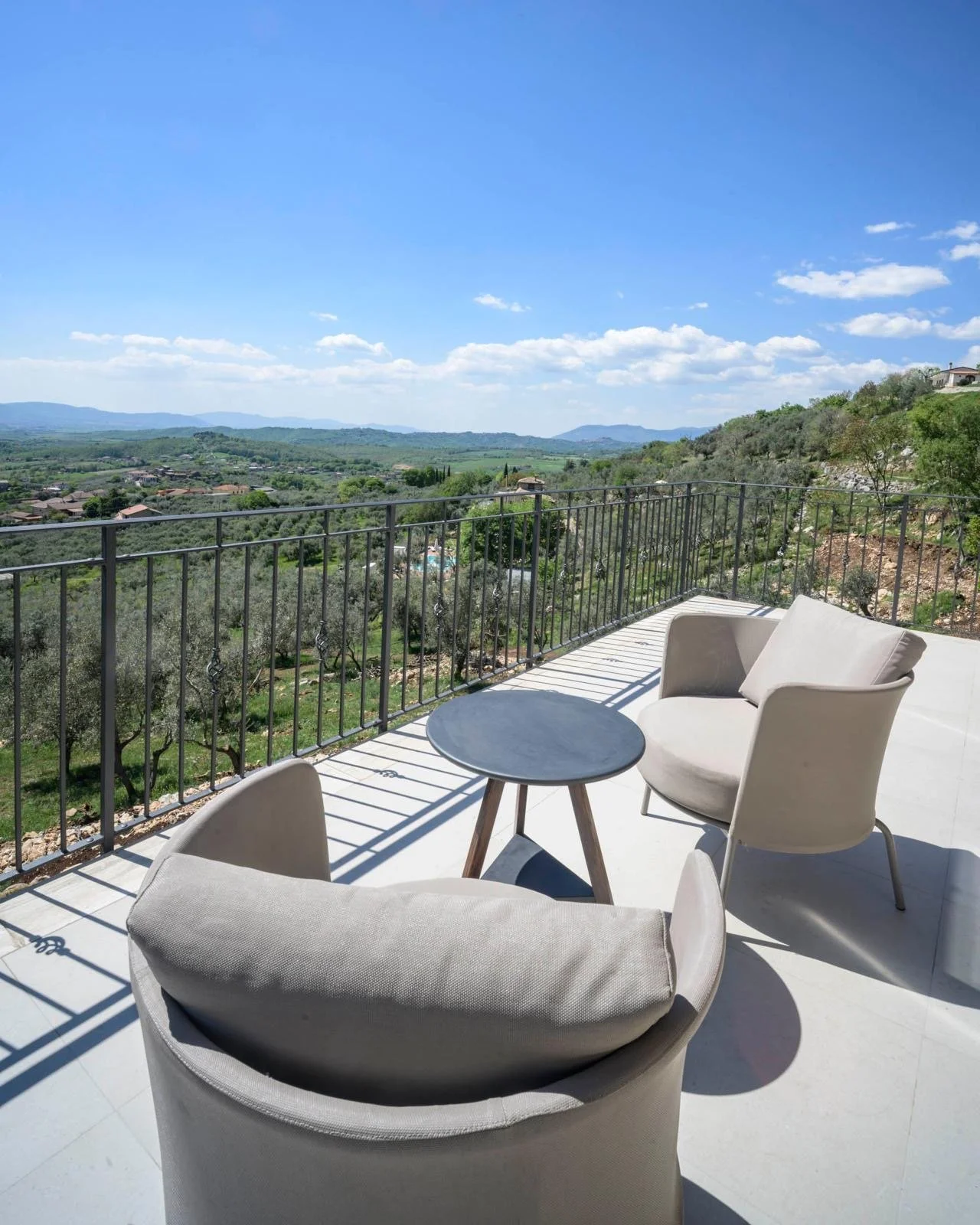 A balcony with modern outdoor furniture, including a rounded armchair with a cushion, another similar armchair, and a small round table, overlooking a scenic landscape of rolling hills, olive trees, and a blue sky with scattered clouds.