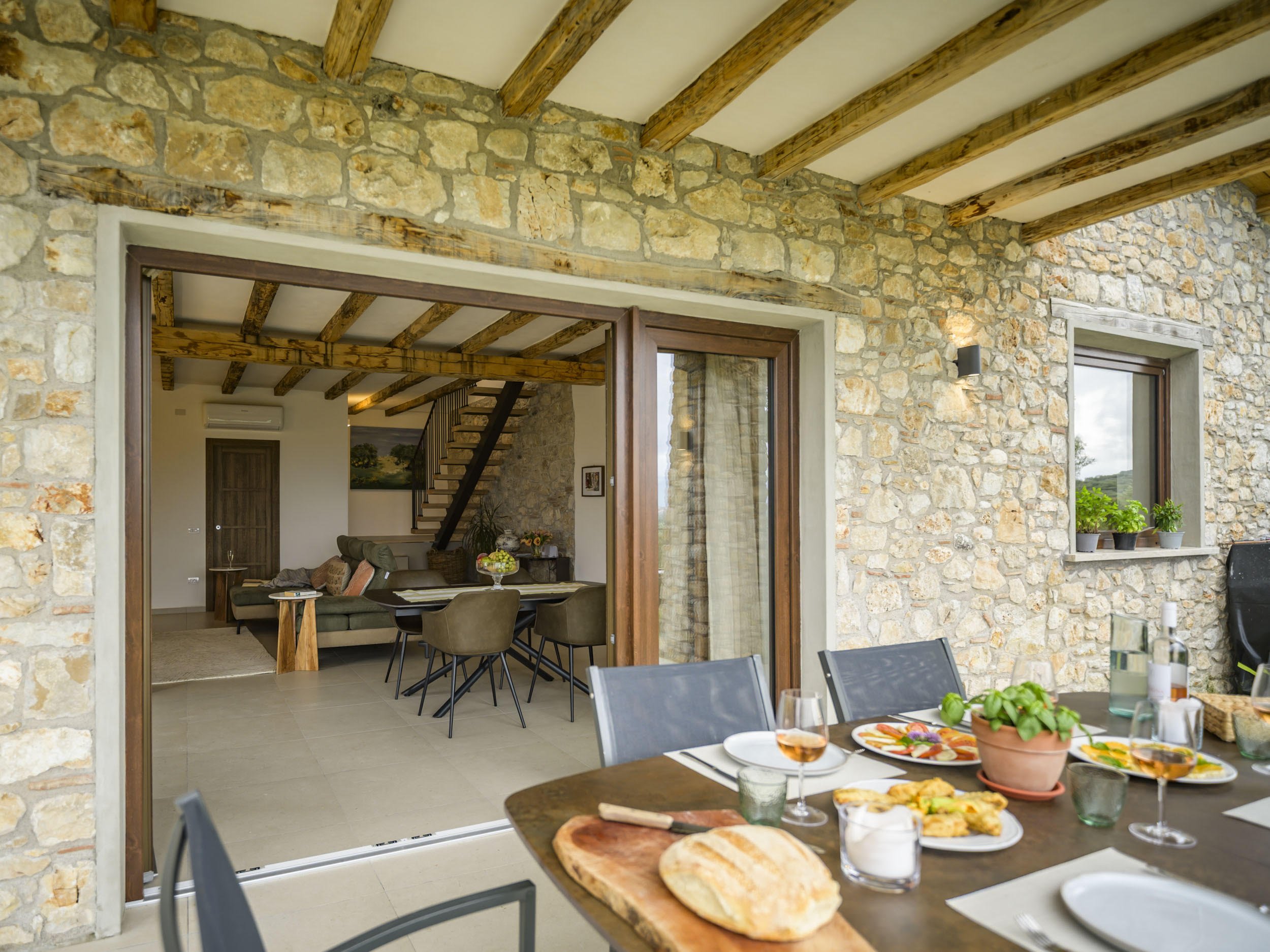Interior of a stone house with wood-beamed ceiling, large windows, and a dining area set for a meal with plates, glasses, and a centerpiece of plants.