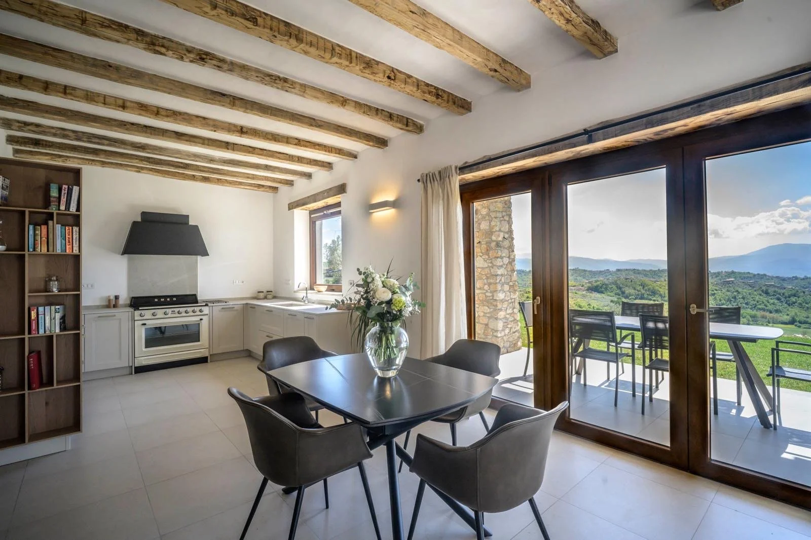 Modern kitchen and dining area with large sliding glass doors overlooking a scenic mountain view, wooden ceiling beams, white cabinetry, black dining table with four chairs, vase with flowers on the table, and an outdoor balcony with seating.