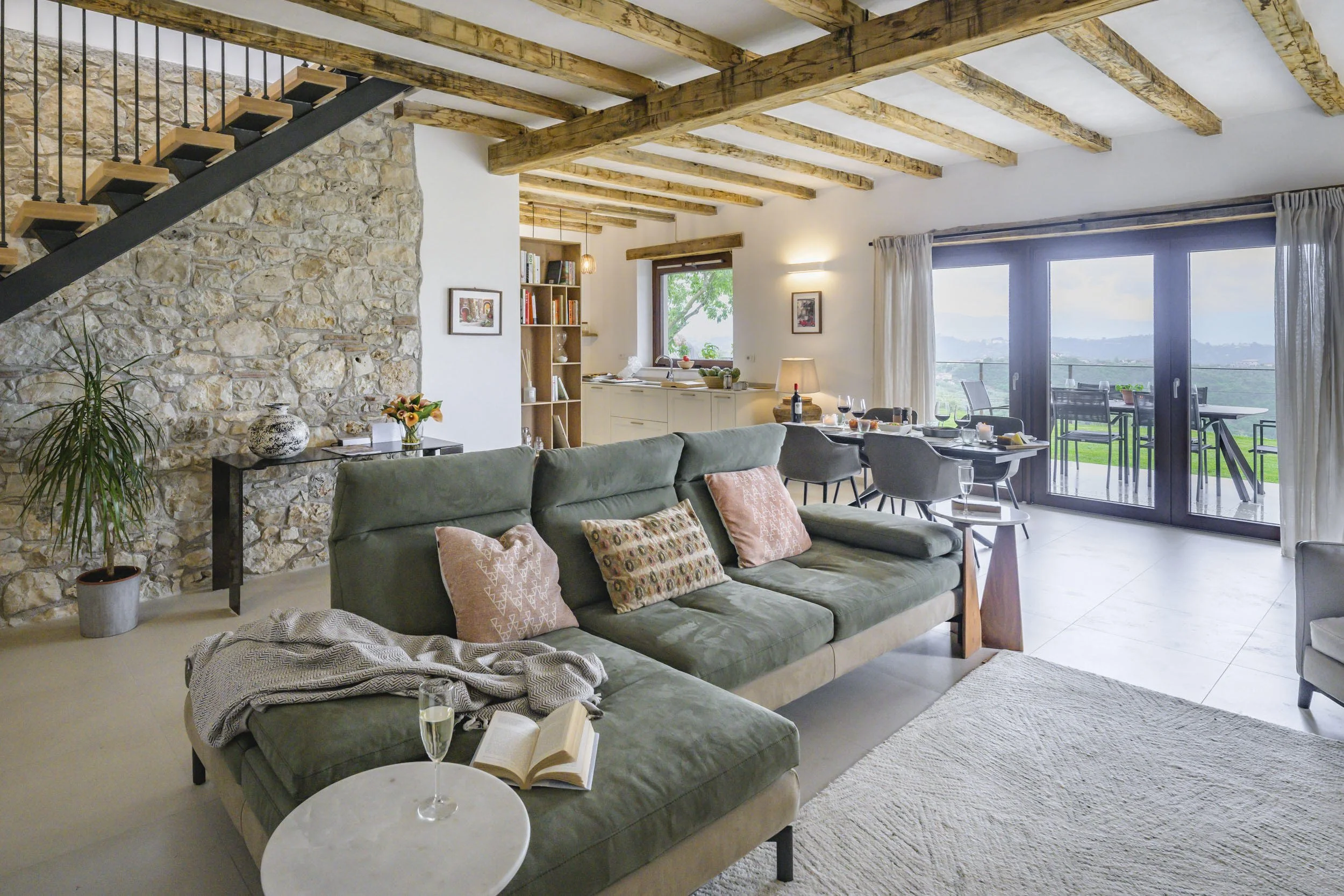 Living room with stone and wooden accents, a large green sofa with patterned pillows, a small white round table with a glass of white wine, an open book, a potted plant, a black console table with a vase and flowers, a wooden bookshelf, and large gla