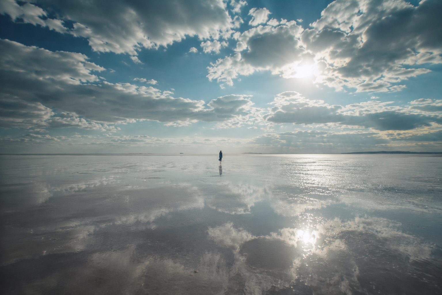 Person standing on a reflective salt flat under a cloudy sky with the sun shining, creating mirror-like reflections of the clouds and sun on the ground.