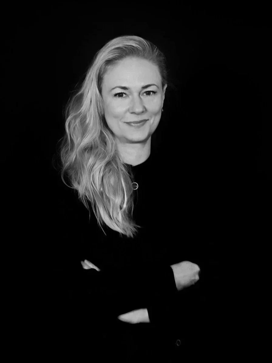 Black and white portrait of a woman with long, wavy hair, smiling, wearing a dark top, against a dark background.