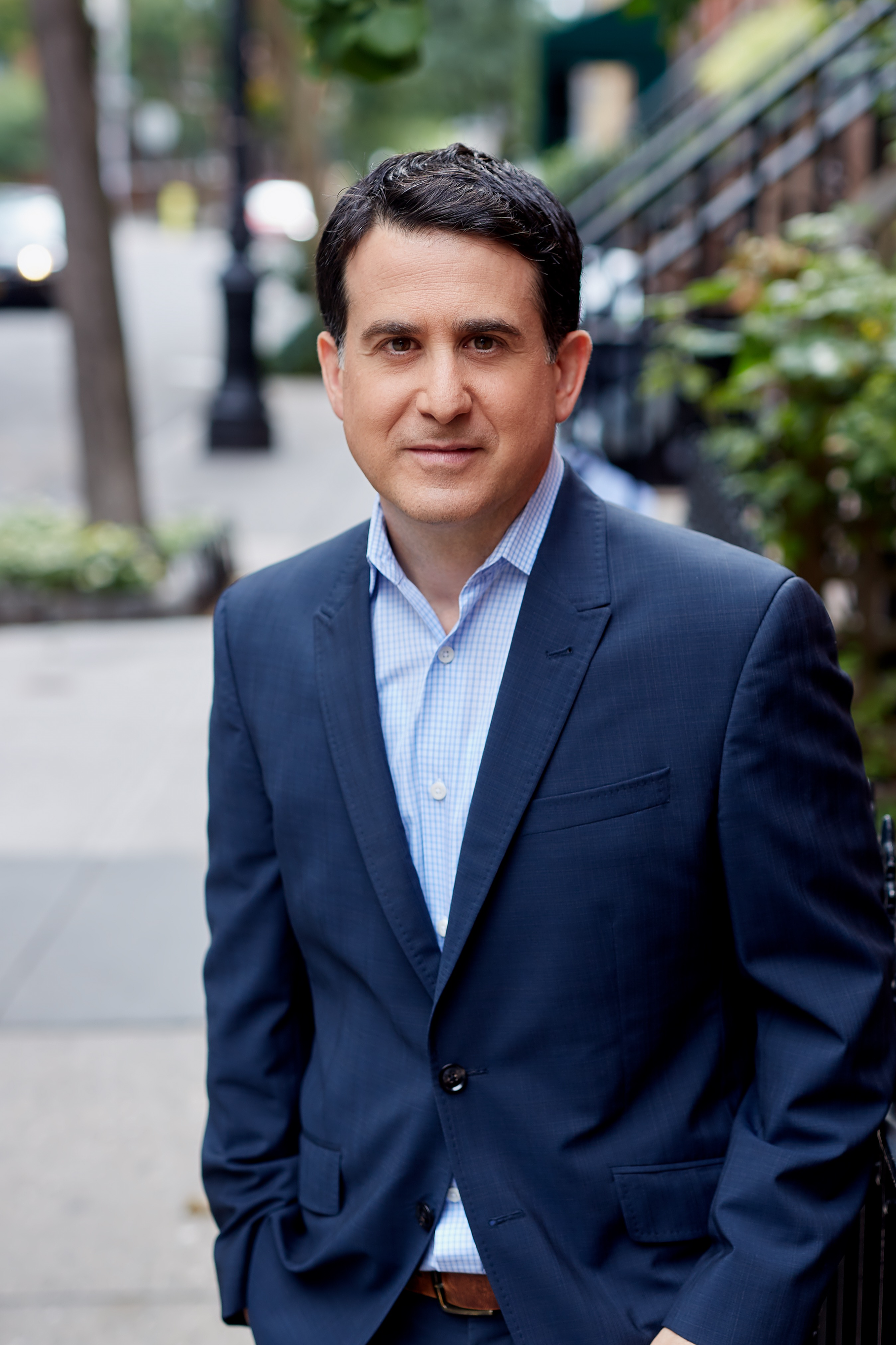 A man in a navy blue suit and light blue dress shirt standing outdoors on a sidewalk, with a tree and stairs in the background.