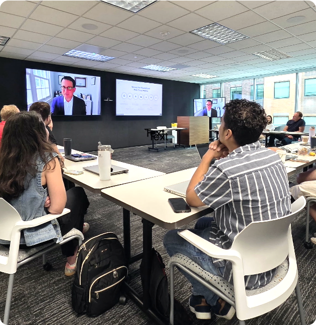 People attending a virtual seminar or conference in a modern conference room with large screens displaying a speaker and presentation slides.