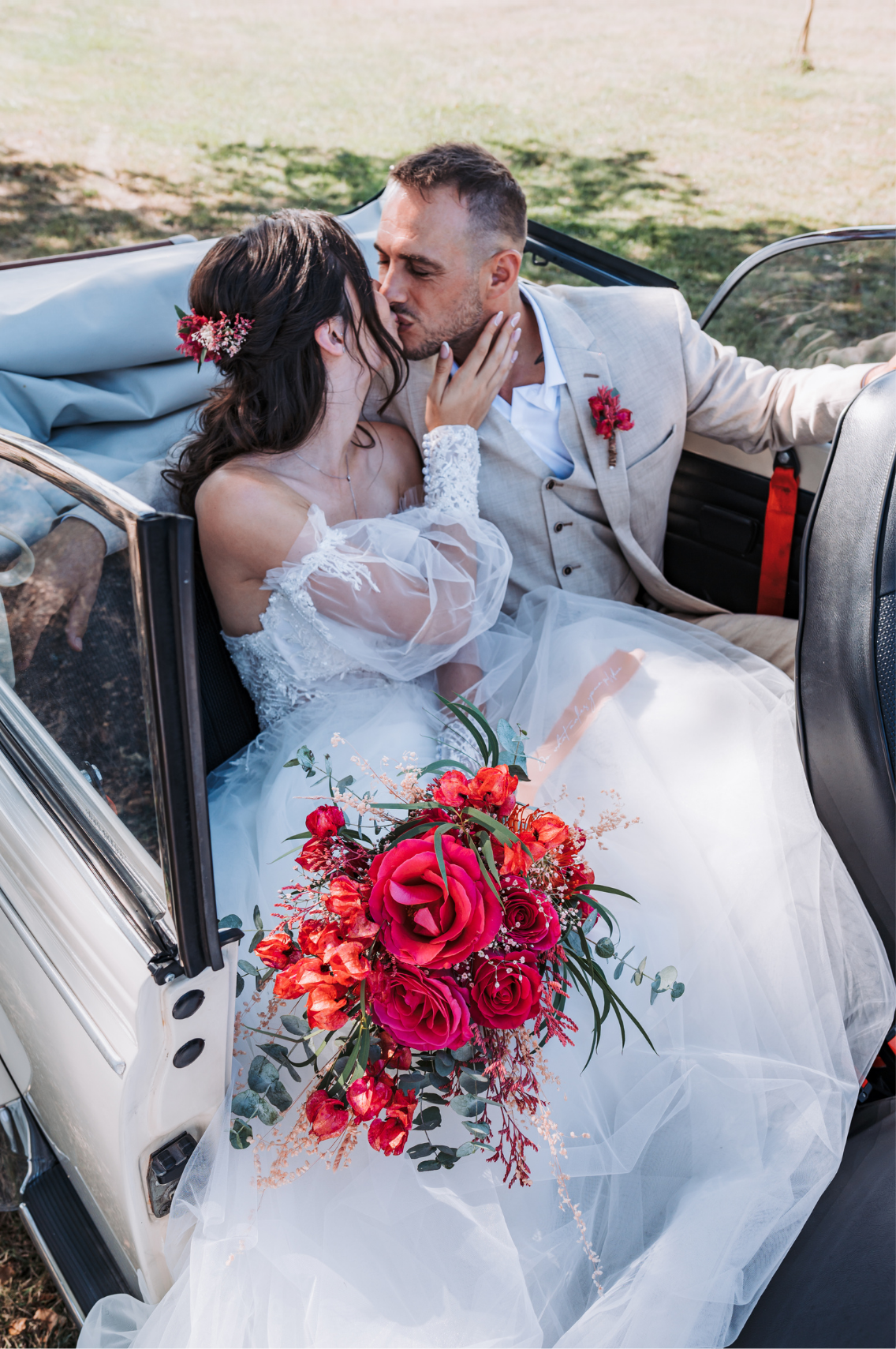 Un couple de mariés s'embrasse dans une voiture décapotable blanche, avec un bouquet de fleurs rouges dans la robe de la mariée.