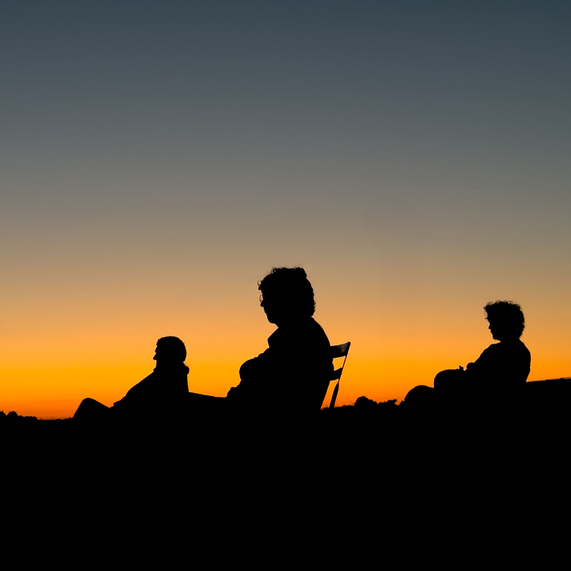 Silhouettes de personnes assises sur le sol lors d'un coucher de soleil, avec un ciel dégradé allant du bleu foncé en haut à l'orange en bas.