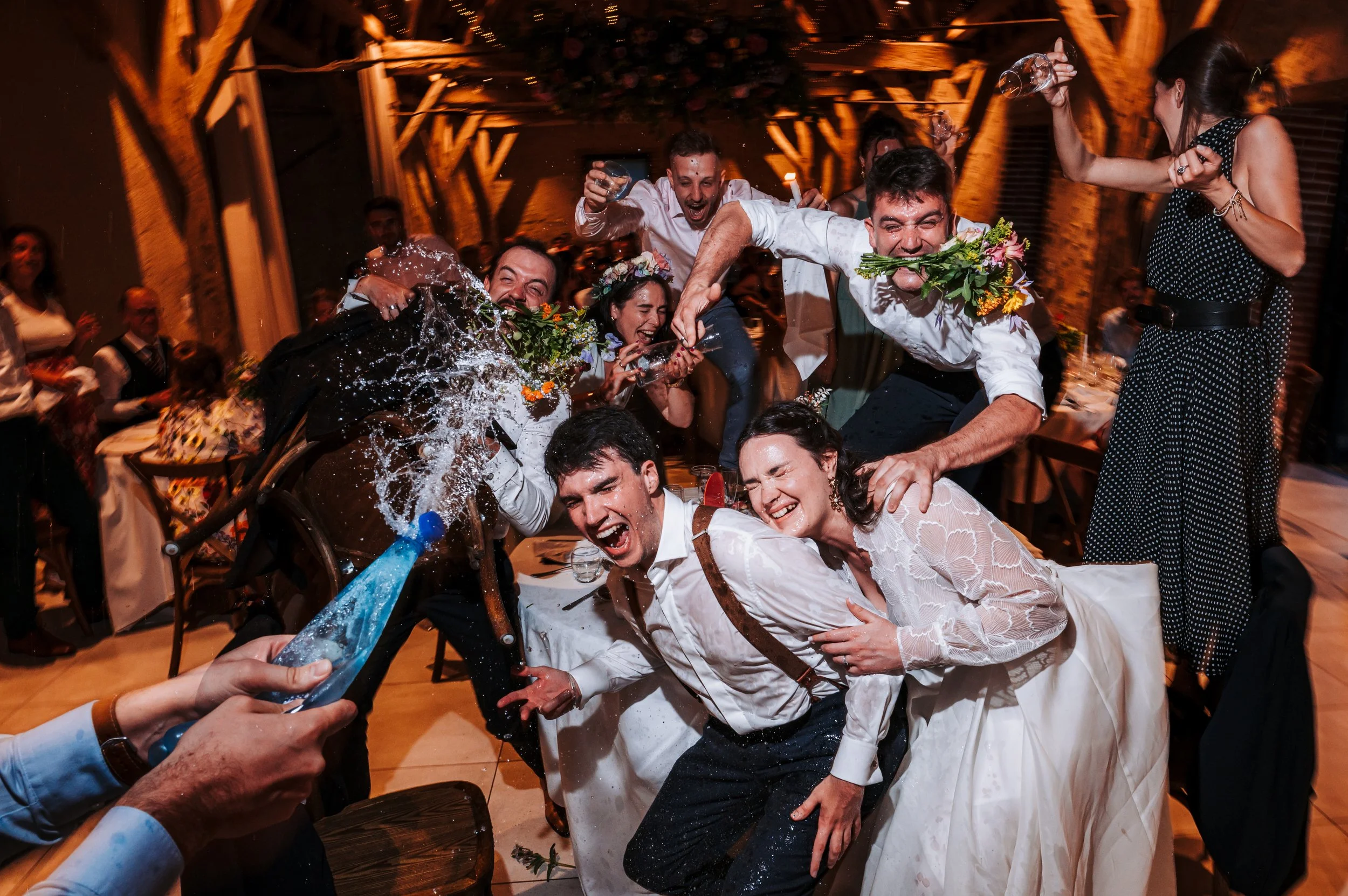 Groupe de personnes célébrant avec des poignées de main et des boissons, le tout dans une ambiance festive à l'intérieur d'un espace décoré. Certains portent des vêtements de mariage.