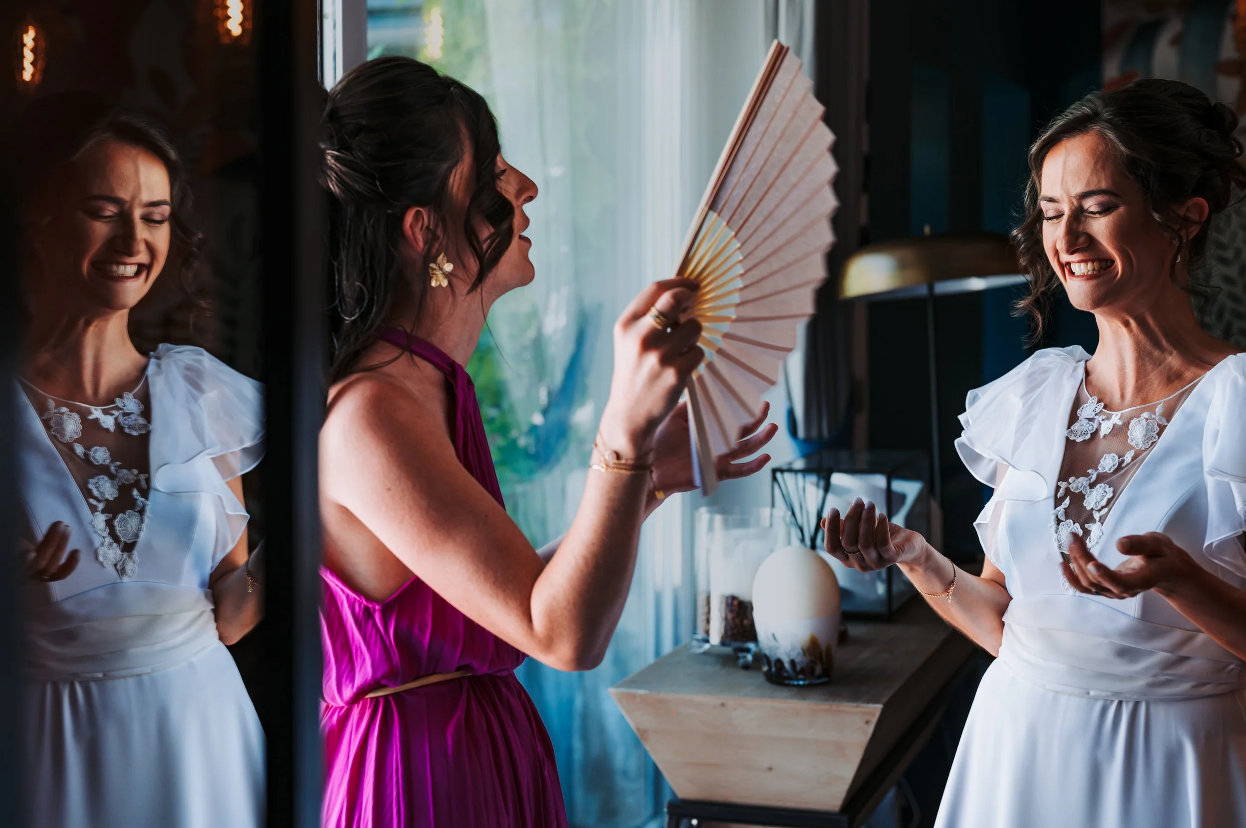 Trois femmes souriantes, dont deux en robes blanches avec broderies, et une en robe rose, discutent et rient dans une cuisine moderne