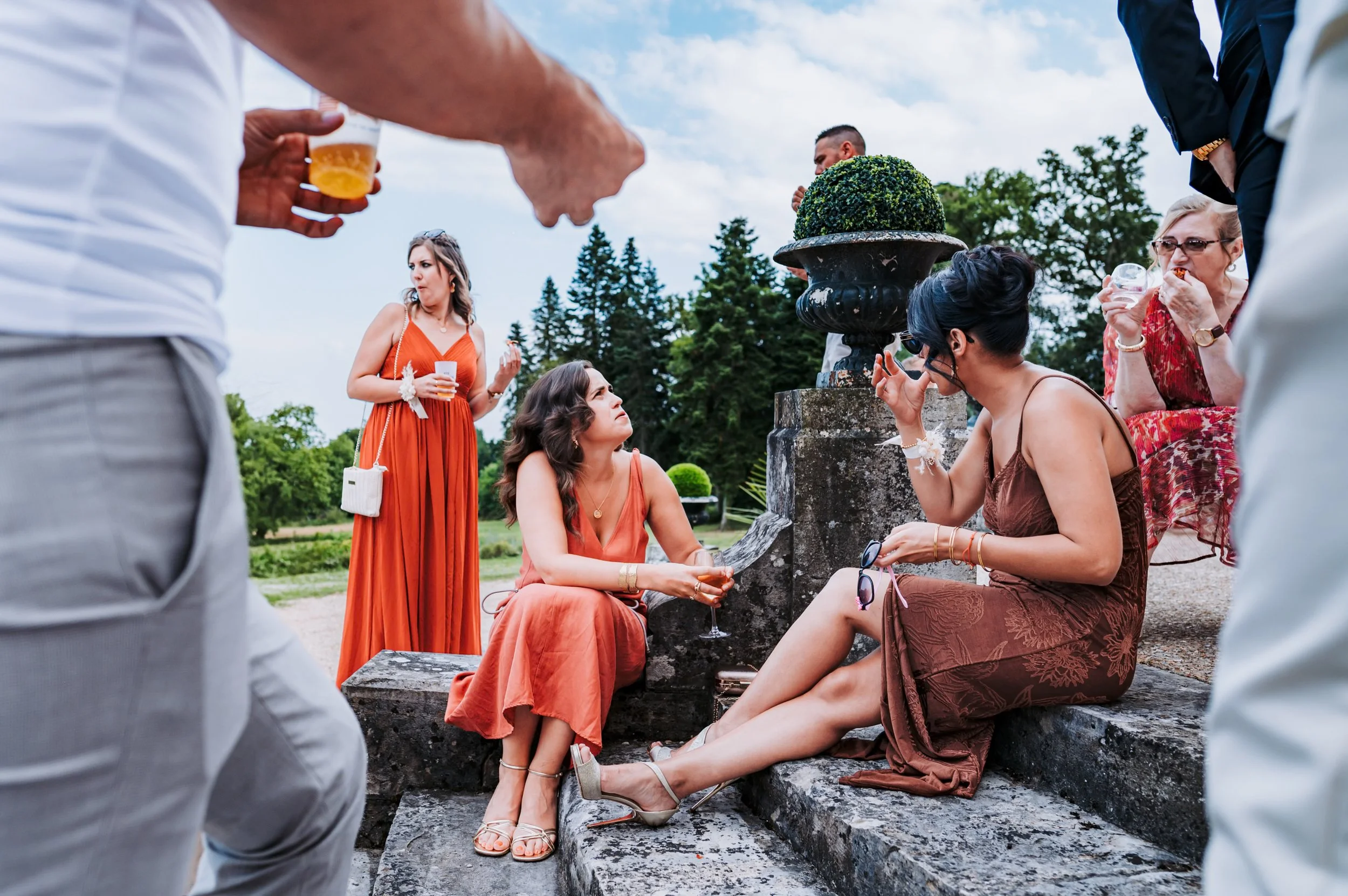 groupe de personnes lors d'une fête en plein air, avec des femmes en robes orange et marron, tenant des verres à vin ou à champagne, discutant et socialisant, dans un jardin avec des arbres en arrière-plan