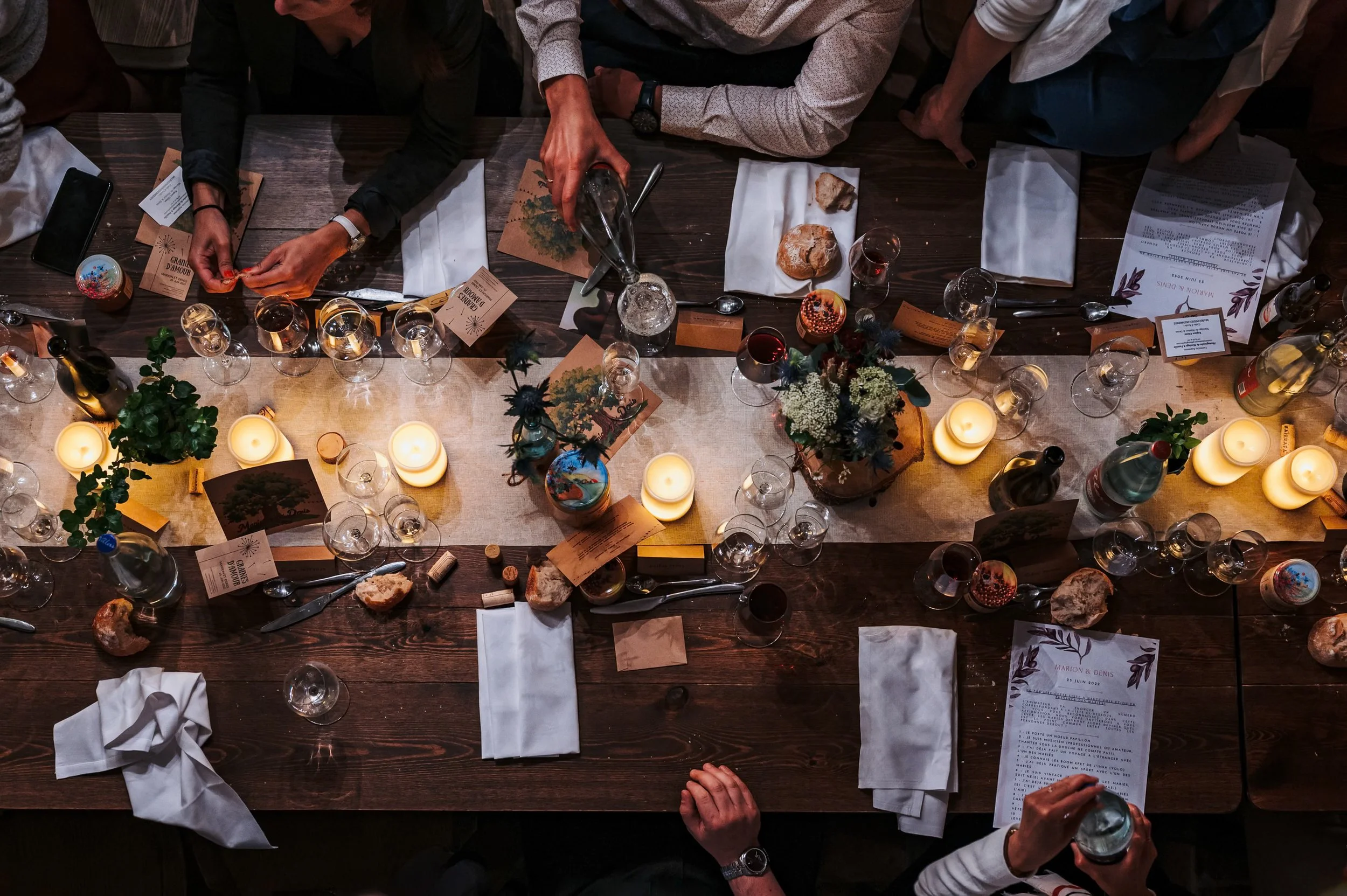 Vue aérienne d'un dîner avec plusieurs personnes autour d'une grande table en bois, entre lesquelles on voit des plats, des verres de vin, des bougies et des décorations florales, dans une ambiance chaleureuse.