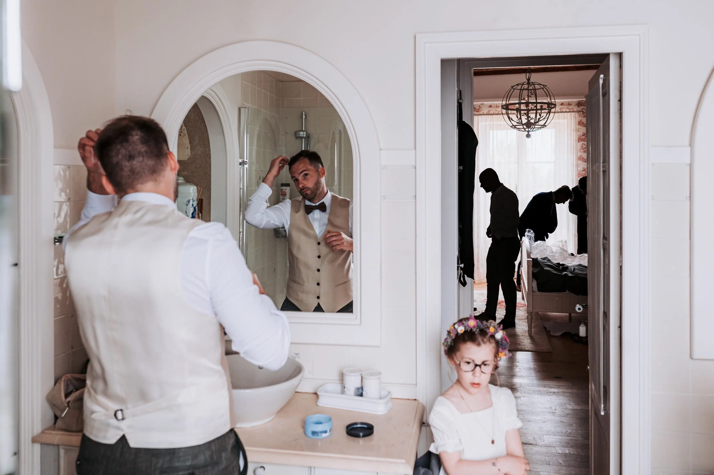 Un homme se coiffe en regardant dans un miroir au-dessus d'un lavabo, tandis qu'un jeune garçon, portant une robe blanche et des lunettes, se tient dans la pièce en face. On voit un autre groupe de personnes dans une autre pièce à travers une porte. L'intérieur est lumineux avec des rideaux et un chandelier suspendu au plafond.