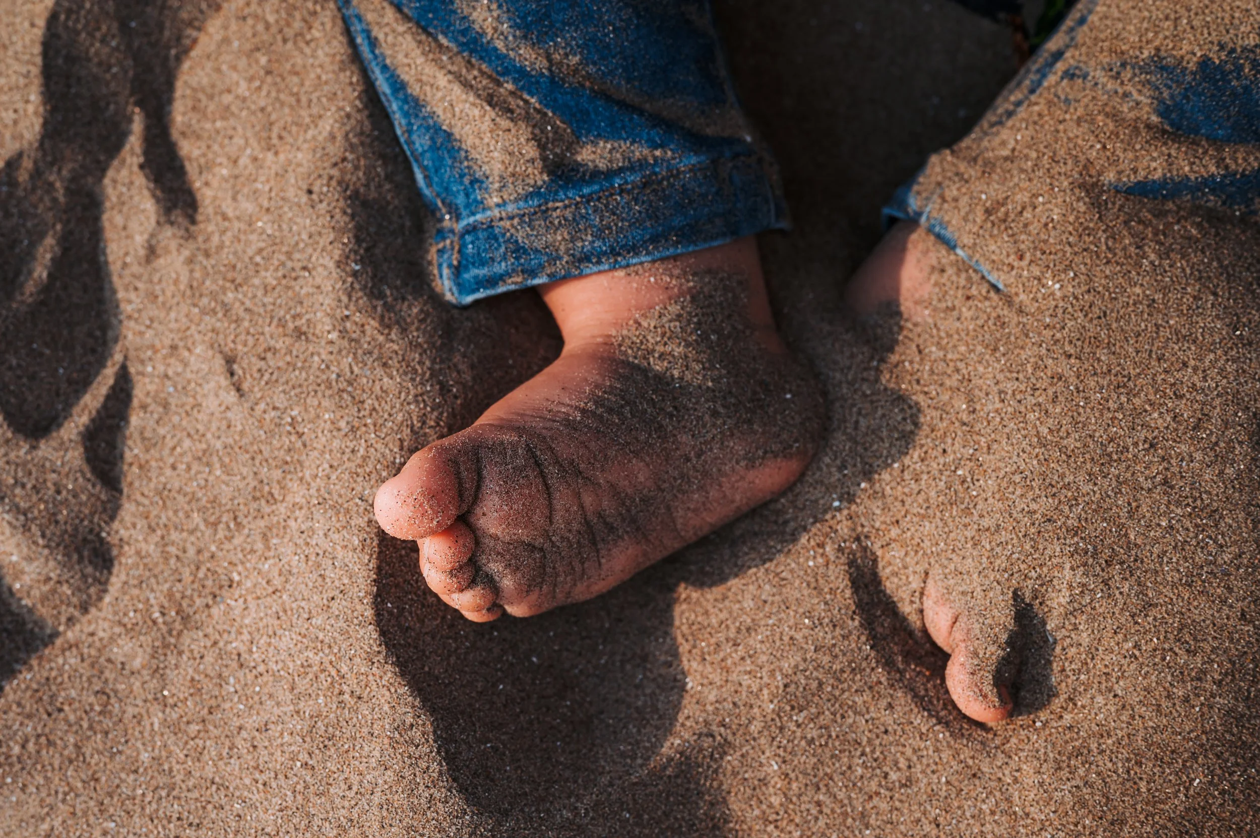 Une main et un pied nus sur du sable, avec des jeans bleus."}**