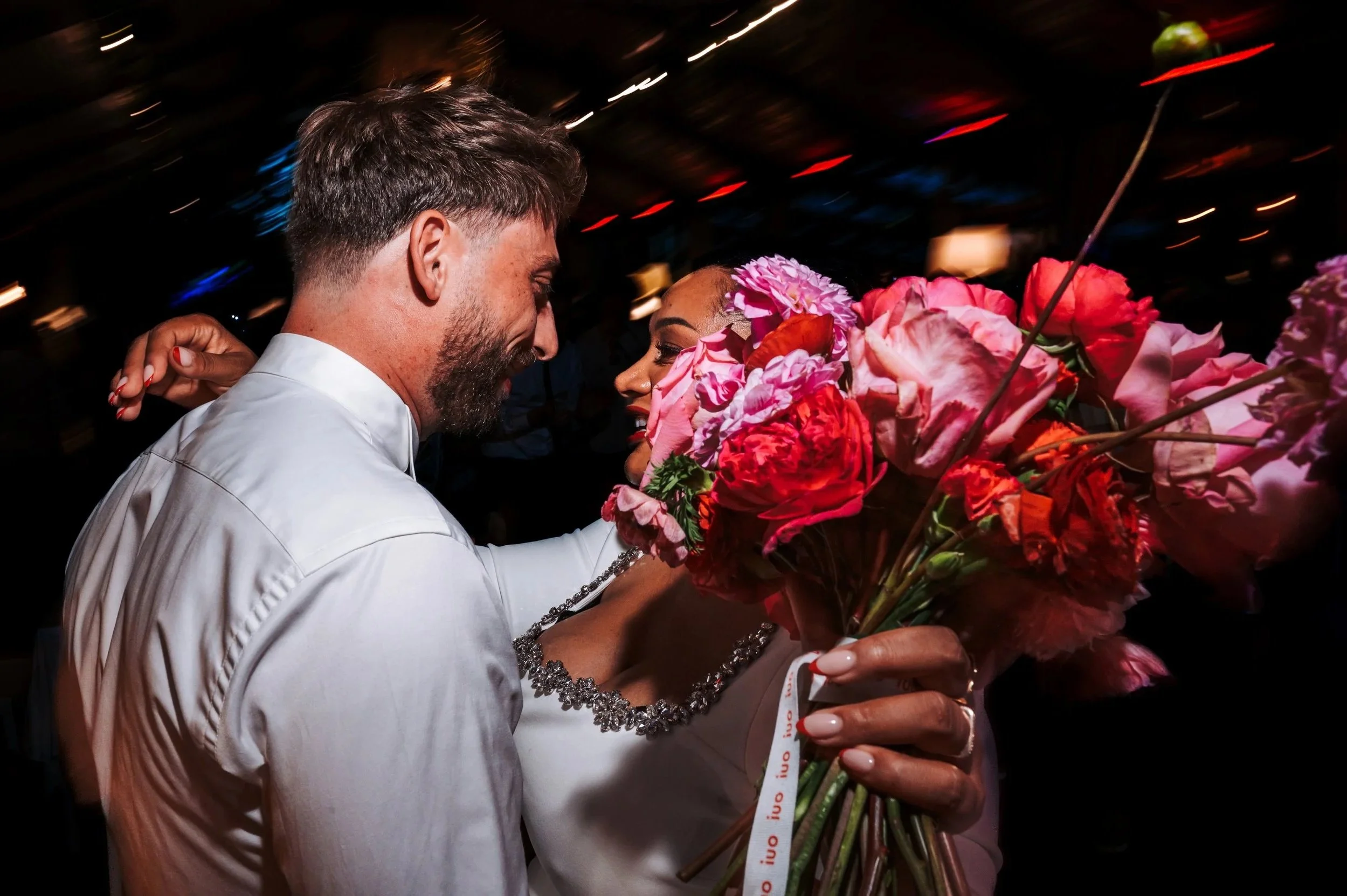 Un homme et une femme s'enlacent, la femme tient un bouquet de fleurs colorées, probablement lors d'une célébration ou d'un événement festif.