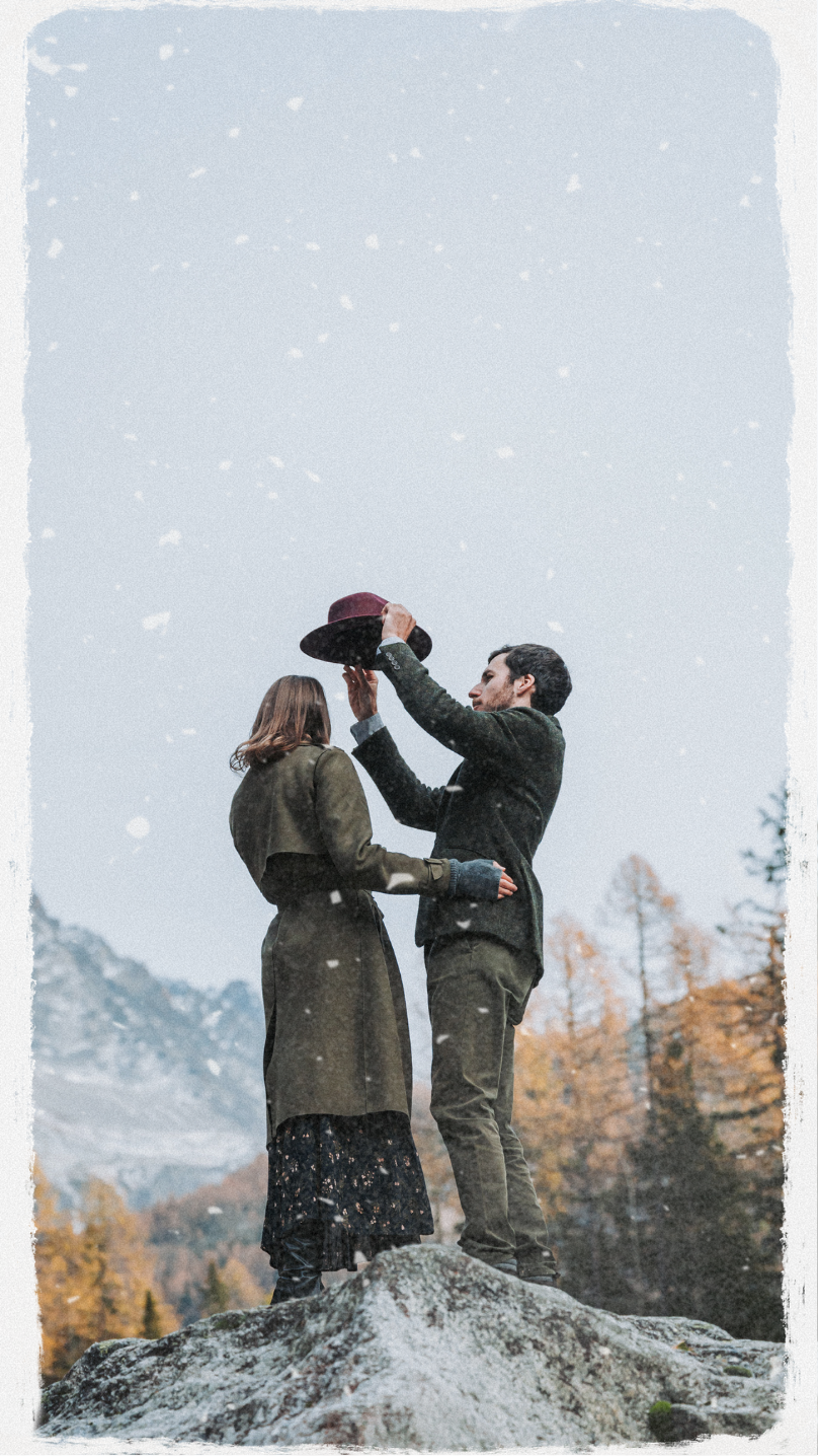 Un homme offre un chapeau à une femme sur un rocher en pleine nature en automne, avec des montagnes enneigées en arrière-plan et des arbres aux feuilles orange.