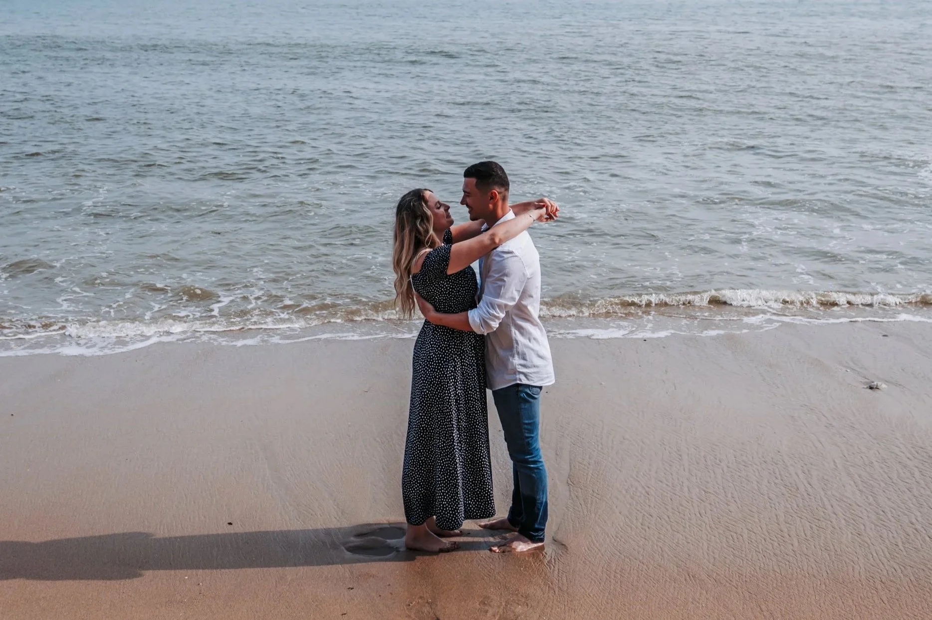 Un couple qui s'aime danse sur la plage au bord de la mer, se regardant dans les yeux et souriant, avec des vagues calmes en arrière-plan.