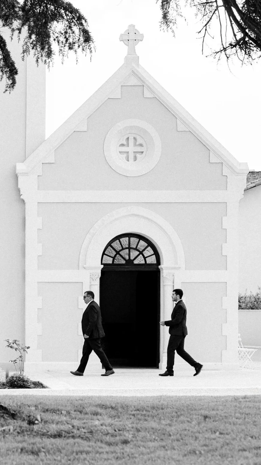 Une petite chapelle blanche avec une croix à son sommet, deux hommes en costumes noirs marchant devant l'entrée, en noir et blanc.