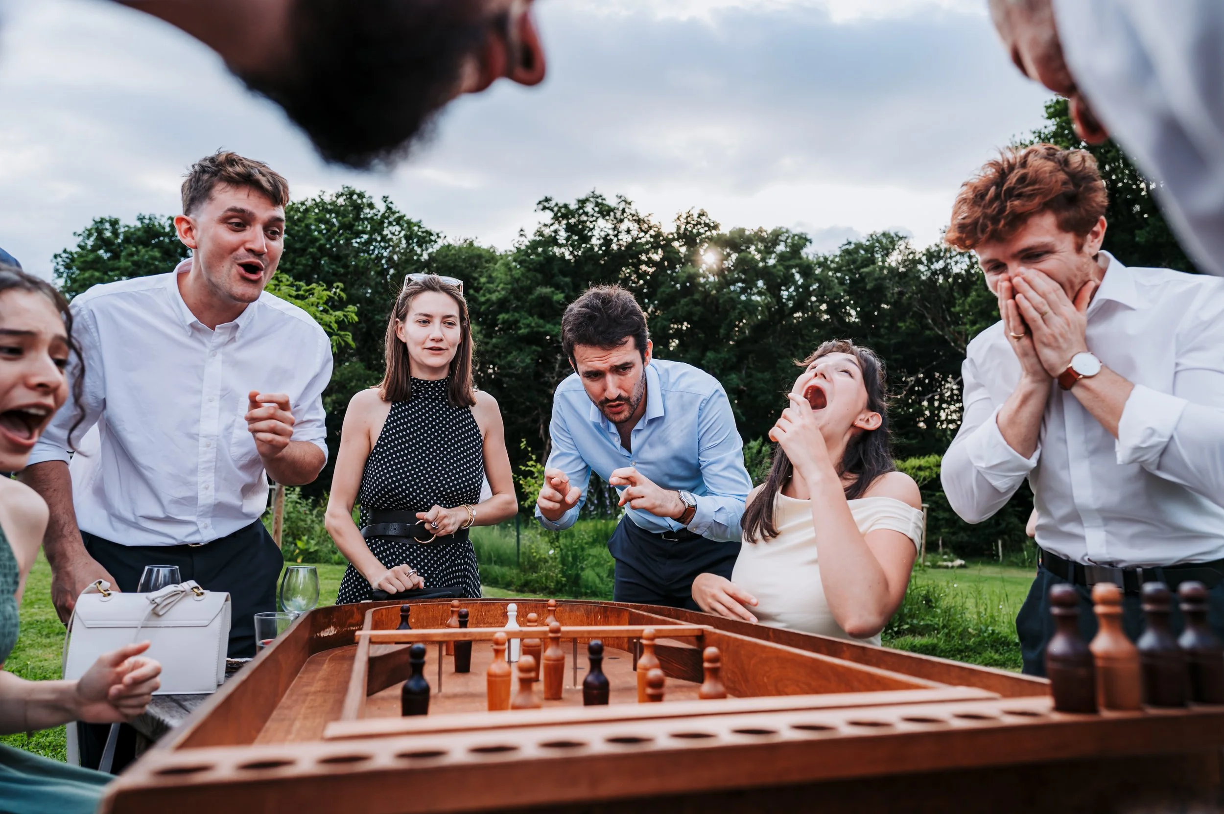 Un groupe de personnes jouent aux échecs en extérieur lors d'une fête ou d'un rassemblement, sous un ciel nuageux, entourées d'arbres et d'une nature verdoyante.