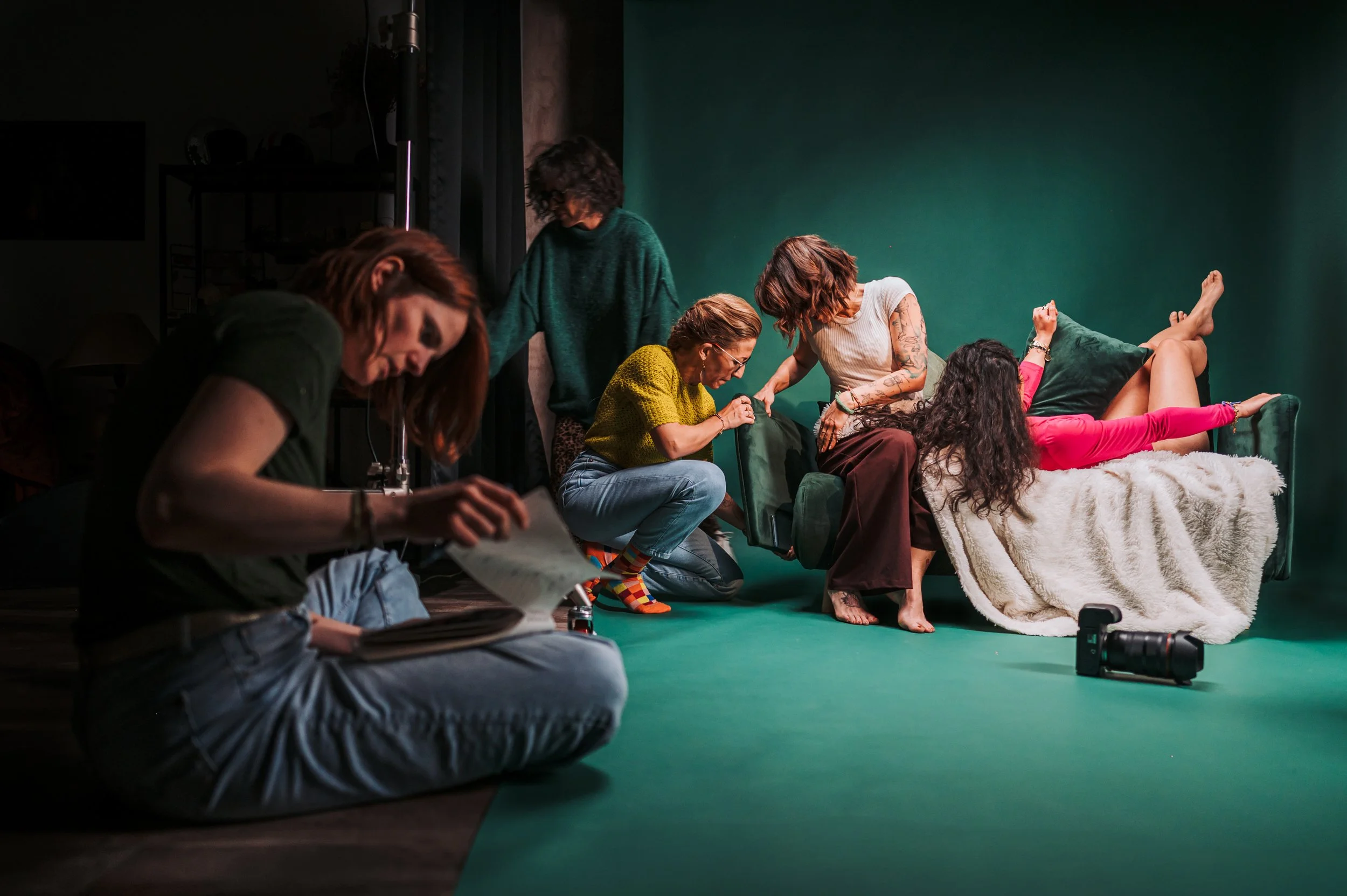 Groupe de personnes en pleine séance de photographie ou de préparation pour une séance photo dans un studio avec un fond vert. Certaines sont assises ou allongées sur un canapé, d'autres ajustent leurs vêtements ou leur posture.