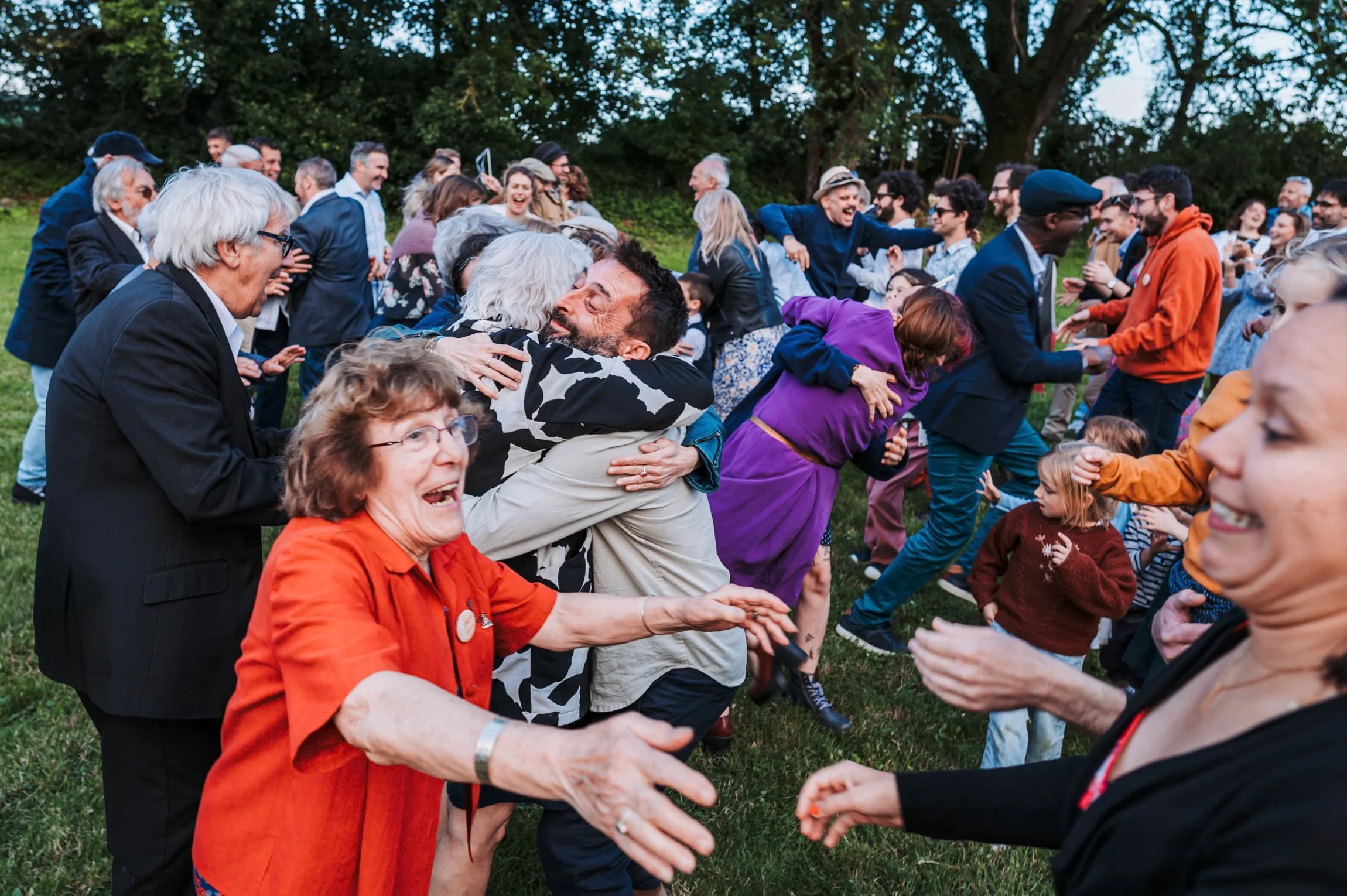 Groupe de personnes souriantes et en train de danser ou de s'amuser lors d'un événement en plein air, avec des hierbes et des arbres à l'arrière-plan.