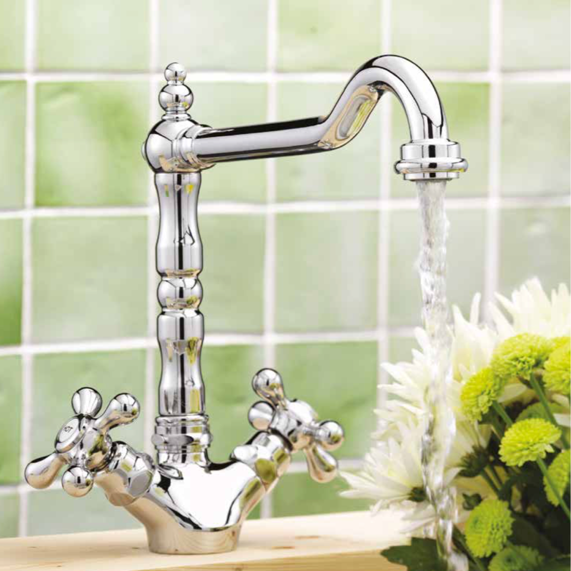 A polished silver kitchen faucet with running water over yellow and white flowers, with a green tile backsplash in the background.