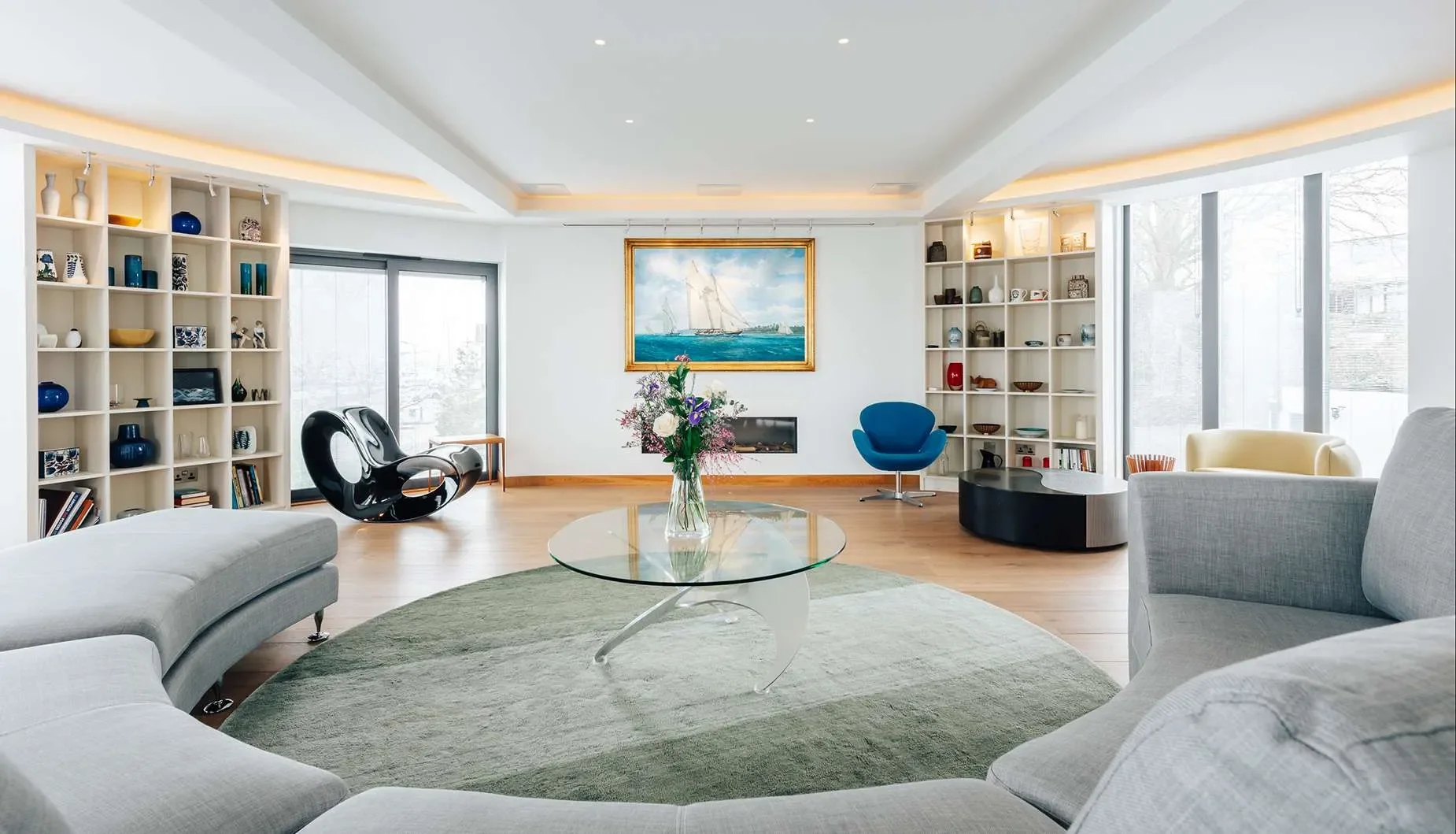 Bright living room with white walls, large windows, and wooden flooring. Features a curved gray sofa around a glass coffee table with a flower vase. Includes a black rounded chair, a blue accent chair, and a yellow armchair. White shelving units with