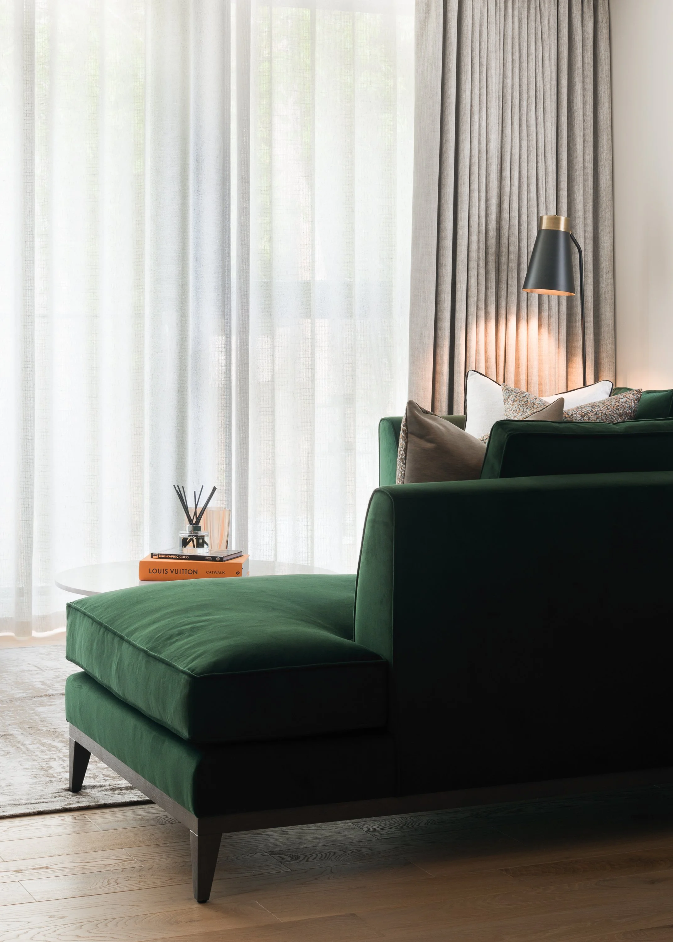 Living room with green velvet sofa, cushions, side table with books, and large window with sheer curtains and gray drapes, illuminated by a standing black and gold lamp.