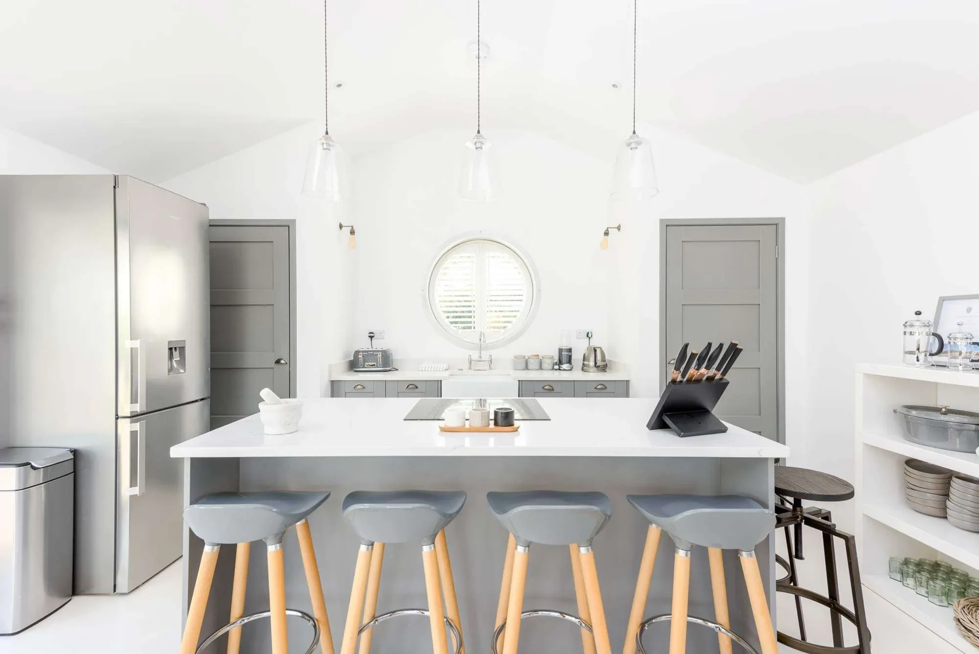 Bright, modern kitchen with white walls and ceiling, gray cabinets, stainless steel refrigerator, and a white island with four light gray stools. The kitchen has pendant lighting, a circular window above the sink, and various appliances and utensils 