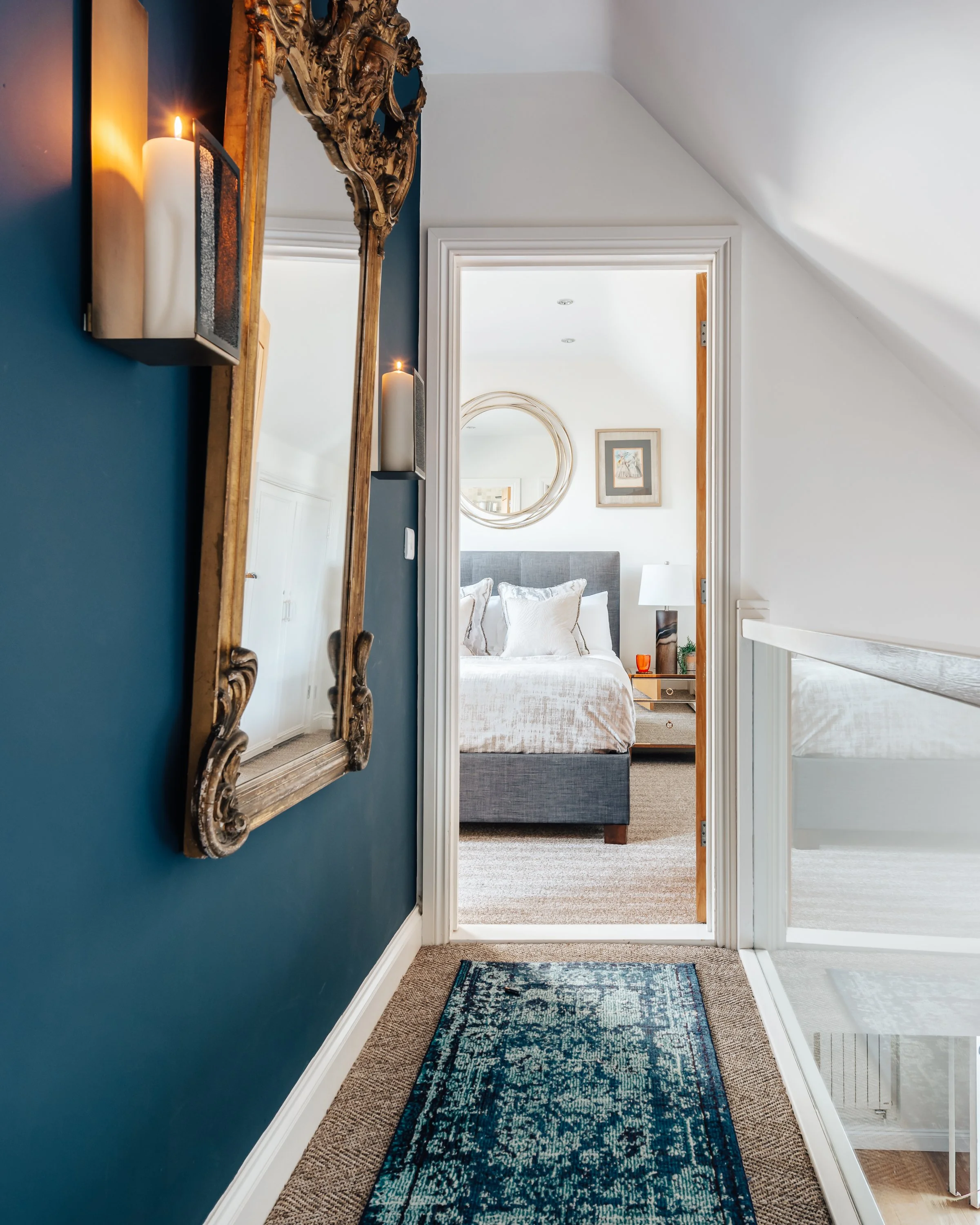 A hallway with a dark blue wall and an ornate wooden mirror, leading into a bedroom. The bedroom features a bed with white bedding, a round mirror on the wall, a framed picture, and a bedside table with an orange glass.