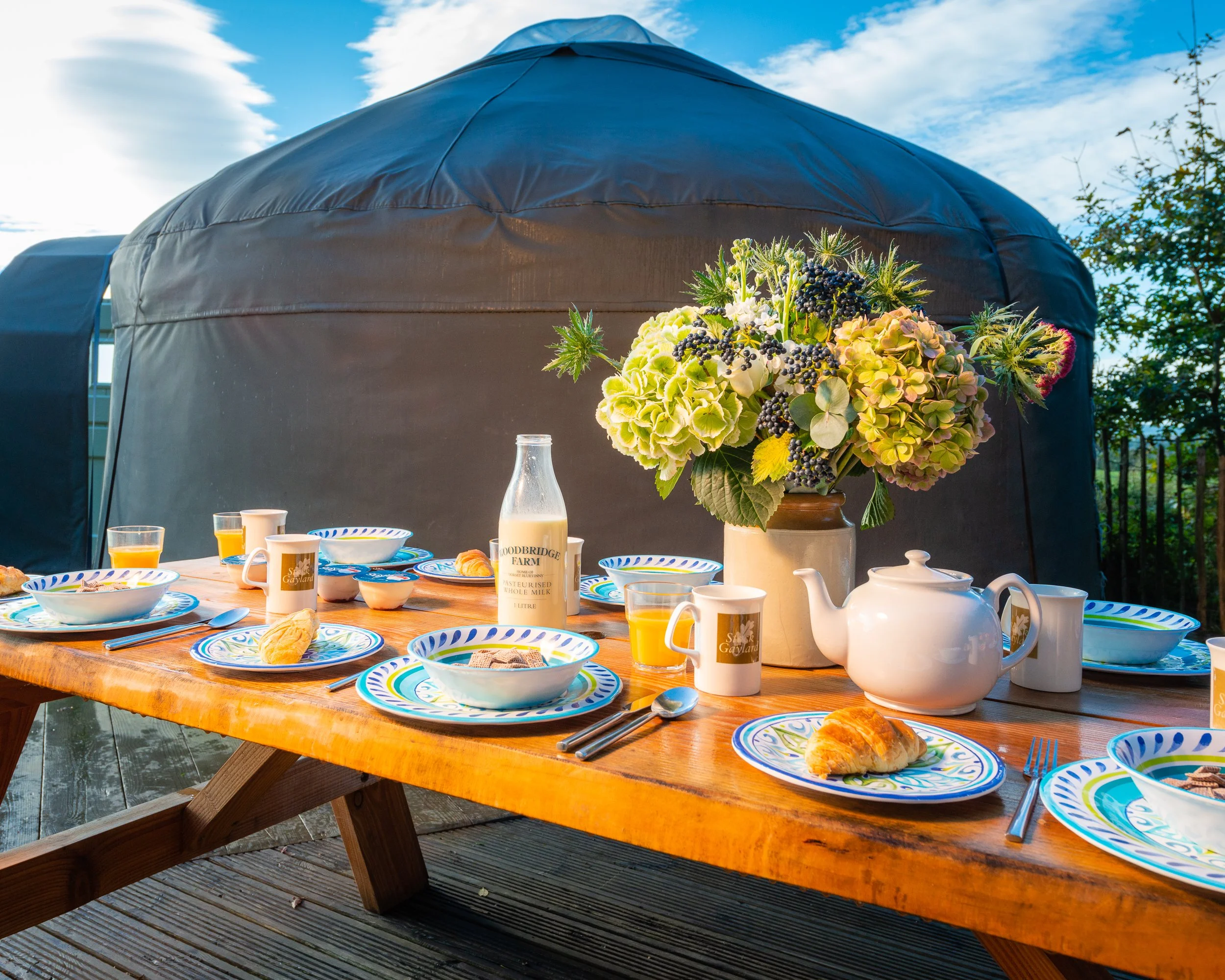 Outdoor breakfast table set beside a glamping yurt at Stock Gaylard, with flowers, crockery and countryside views.