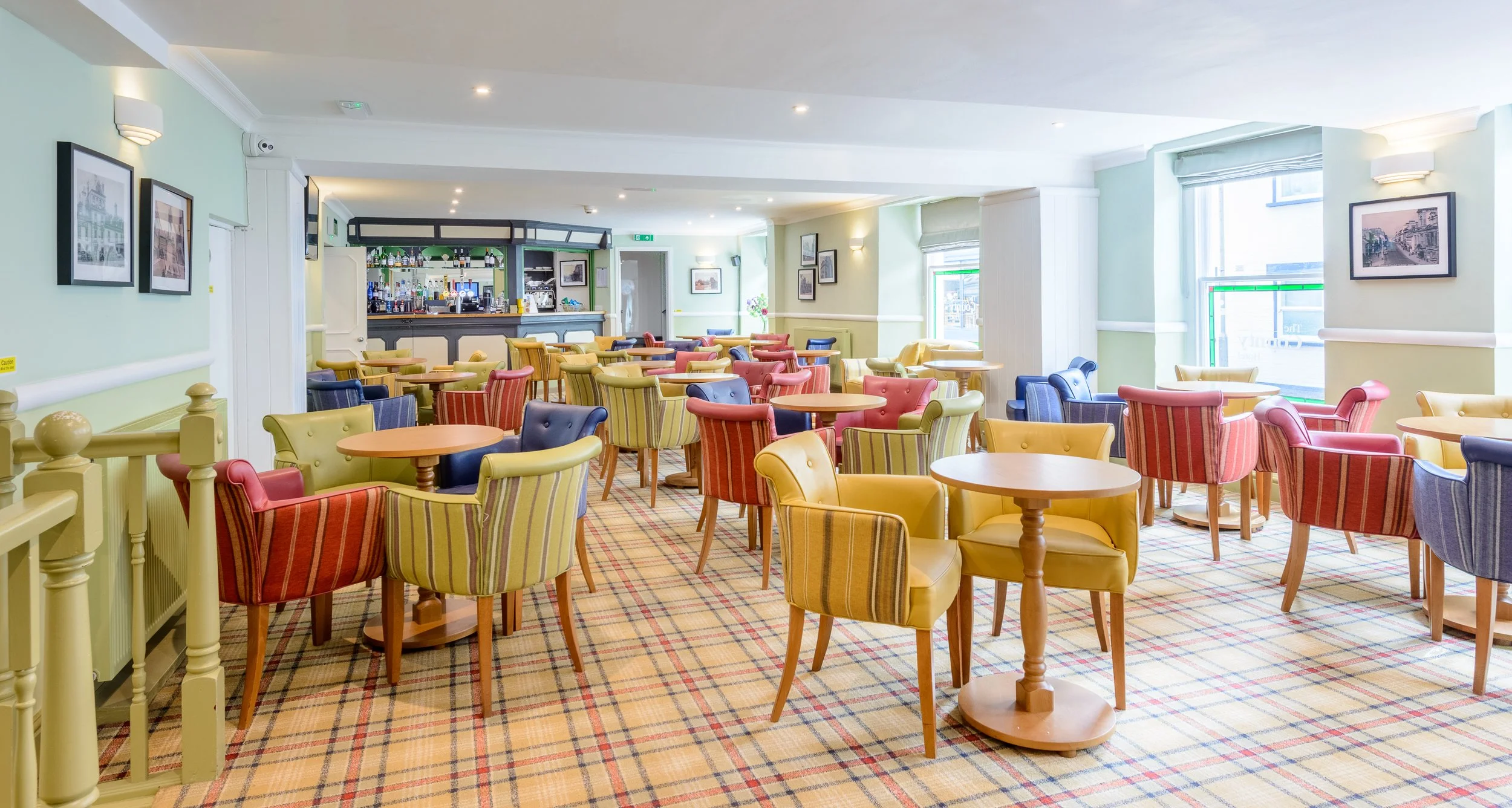 Colorful seating area in a restaurant or café with yellow, pink, blue, and striped armchairs around round wooden tables. Light green walls with framed pictures, windows letting in natural light, and a bar area in the background.