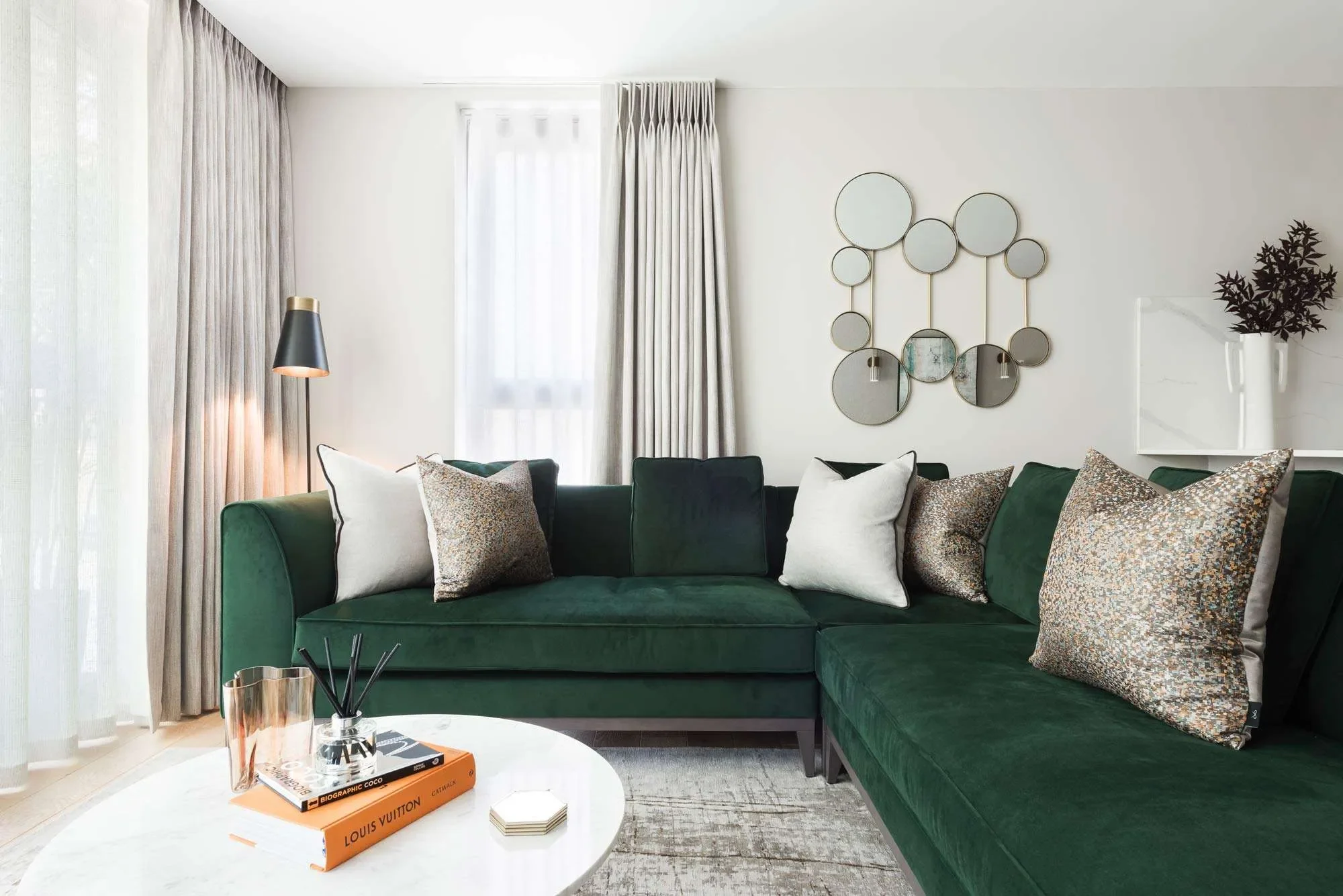 Modern living room with green velvet sectional sofa, patterned throw pillows, a white marble coffee table with books and decorative items, wall mirror art, a tall floor lamp, and large windows with sheer curtains.