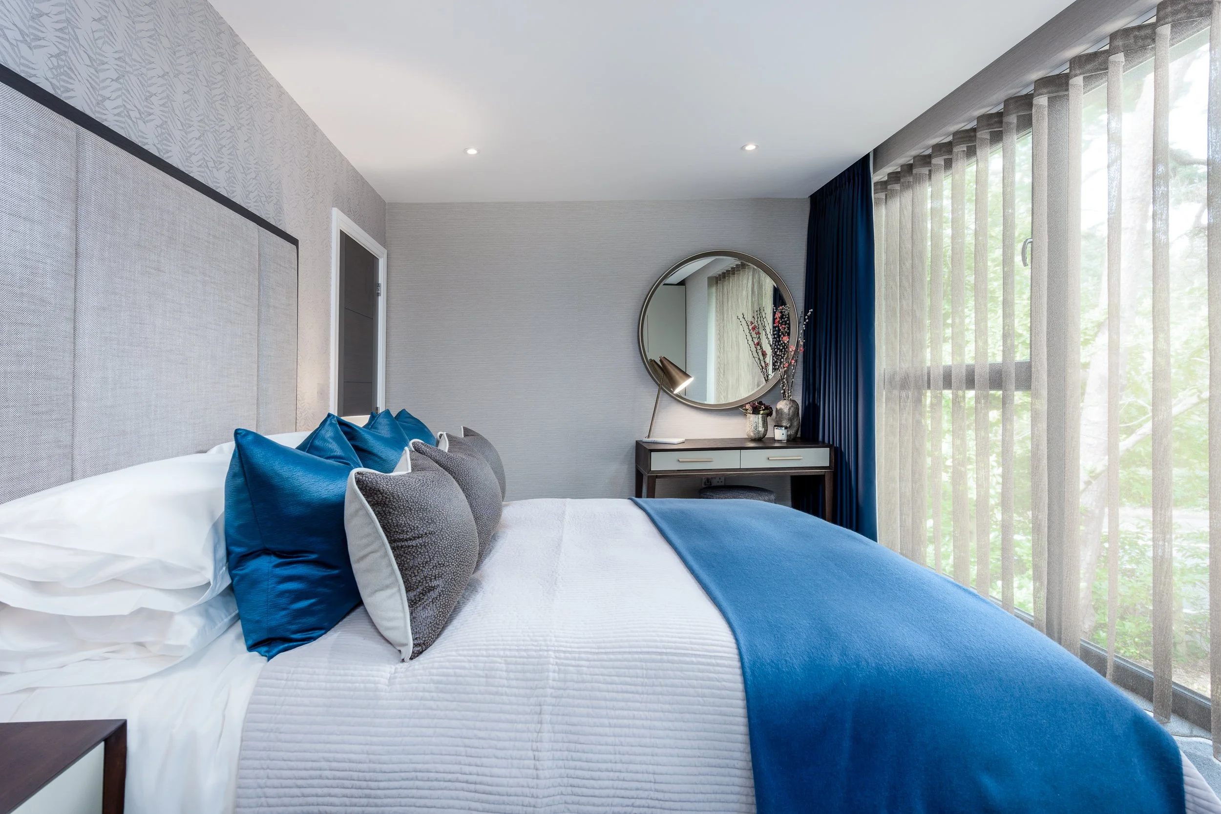 Contemporary bedroom interior featuring blue accent cushions, upholstered headboard, dressing table, round mirror and full-height window treatments.