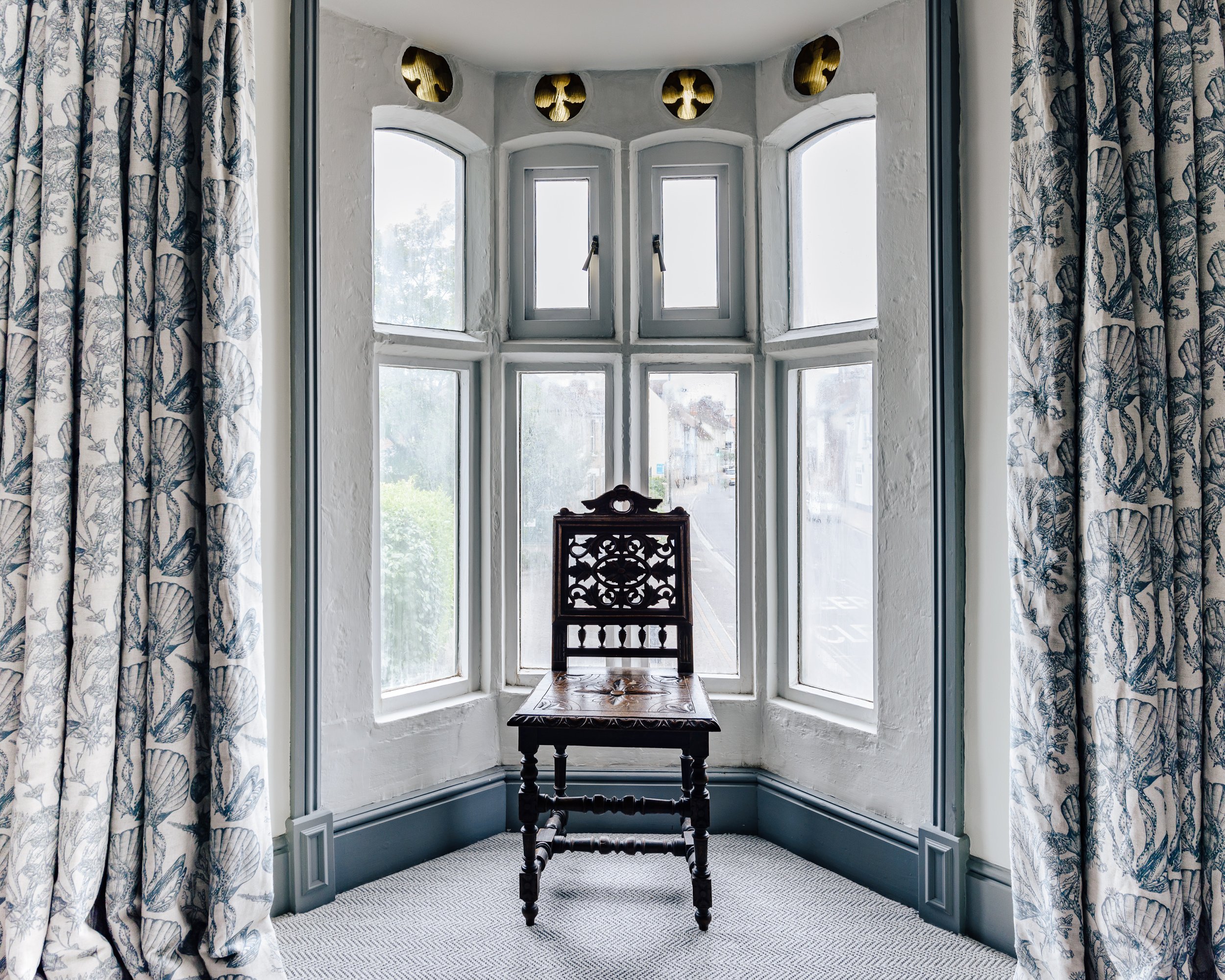 A window seat with a dark wood chair placed in front of a multi-pane window setup with white walls and blue molding. The window features decorative round vents above. There are long patterned curtains on either side of the window, and the floor is carpeted in a light grey and white geometric pattern.