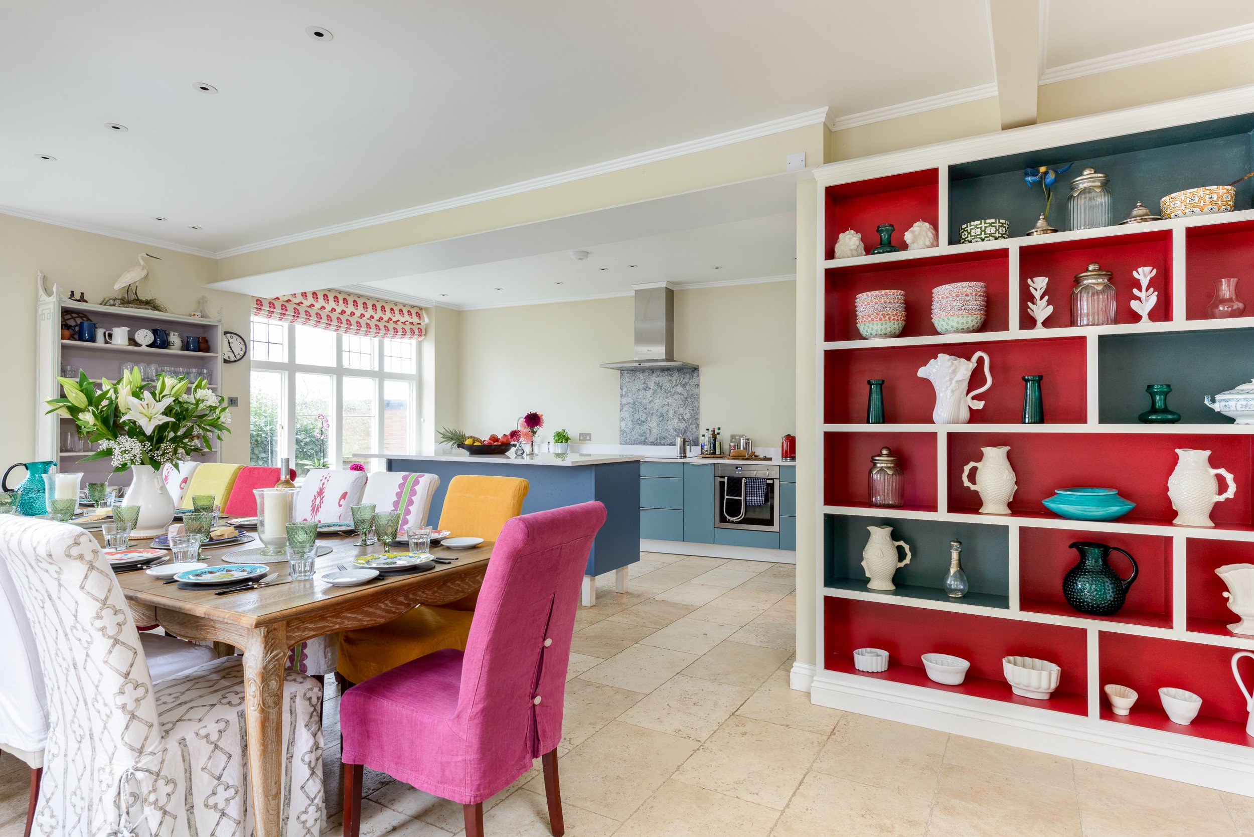 Kitchen and dining room at The Pink House, showing a large dining table, colourful chairs, blue kitchen cabinetry and red display shelving.