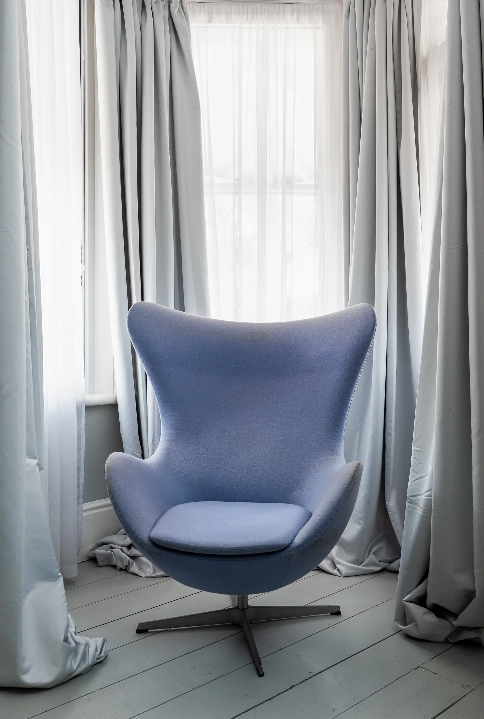 A light blue modern swivel chair with a curved backrest, positioned in front of a window with white curtains. The curtains are partially drawn, allowing light to fill the room, and the floor is made of light-colored wooden planks.