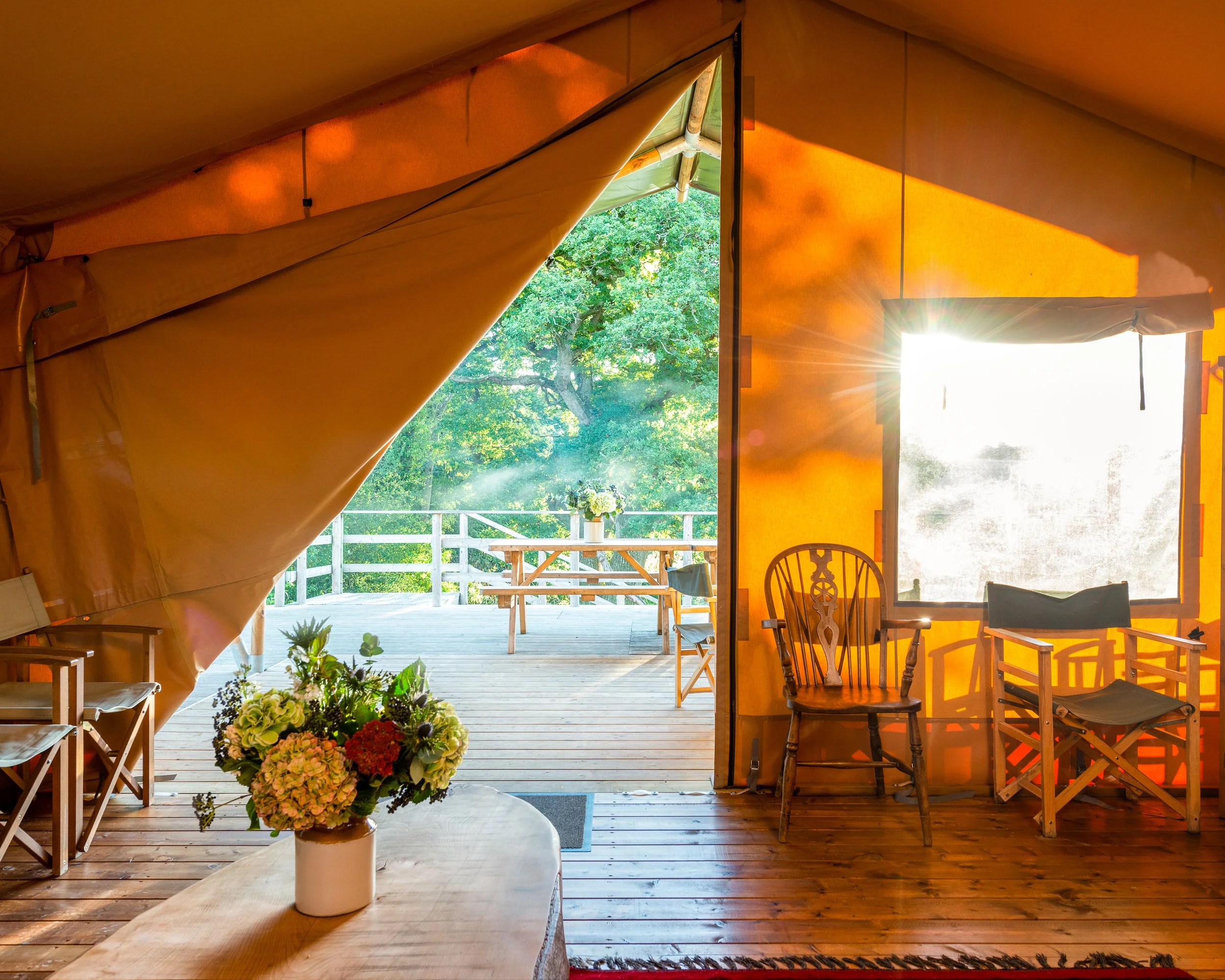 Interior view of a cozy tent with a view of green trees outside. The tent has orange fabric walls, wooden furniture, and a table with a vase of flowers. Sunlight streams in through an open door leading to an outdoor deck with a picnic table.