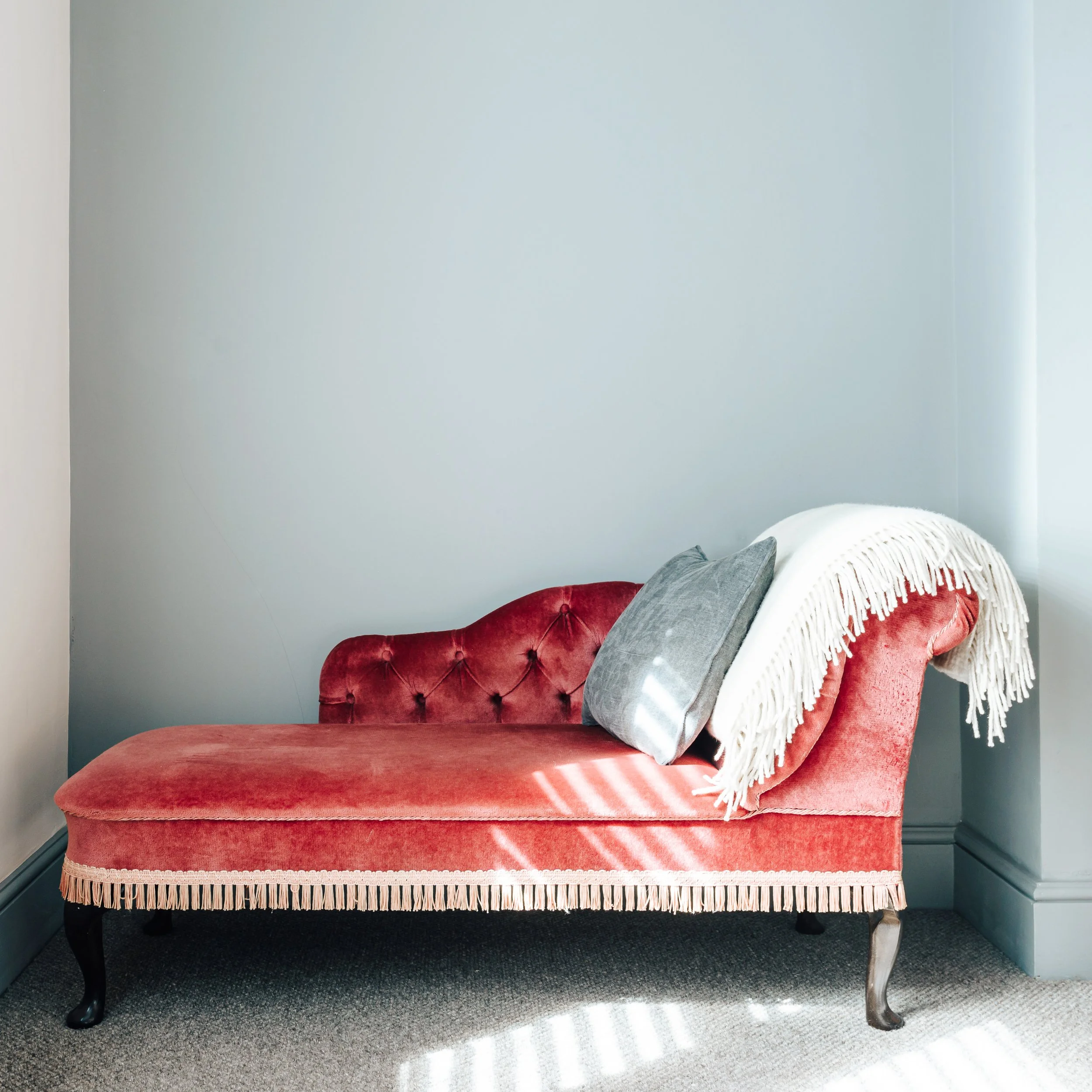 A vintage red upholstered settee with black wooden legs, topped with gray and white pillows, and a white fringed throw blanket. The settee is placed against a light gray wall, with sunlight casting shadows on the gray carpeted floor.