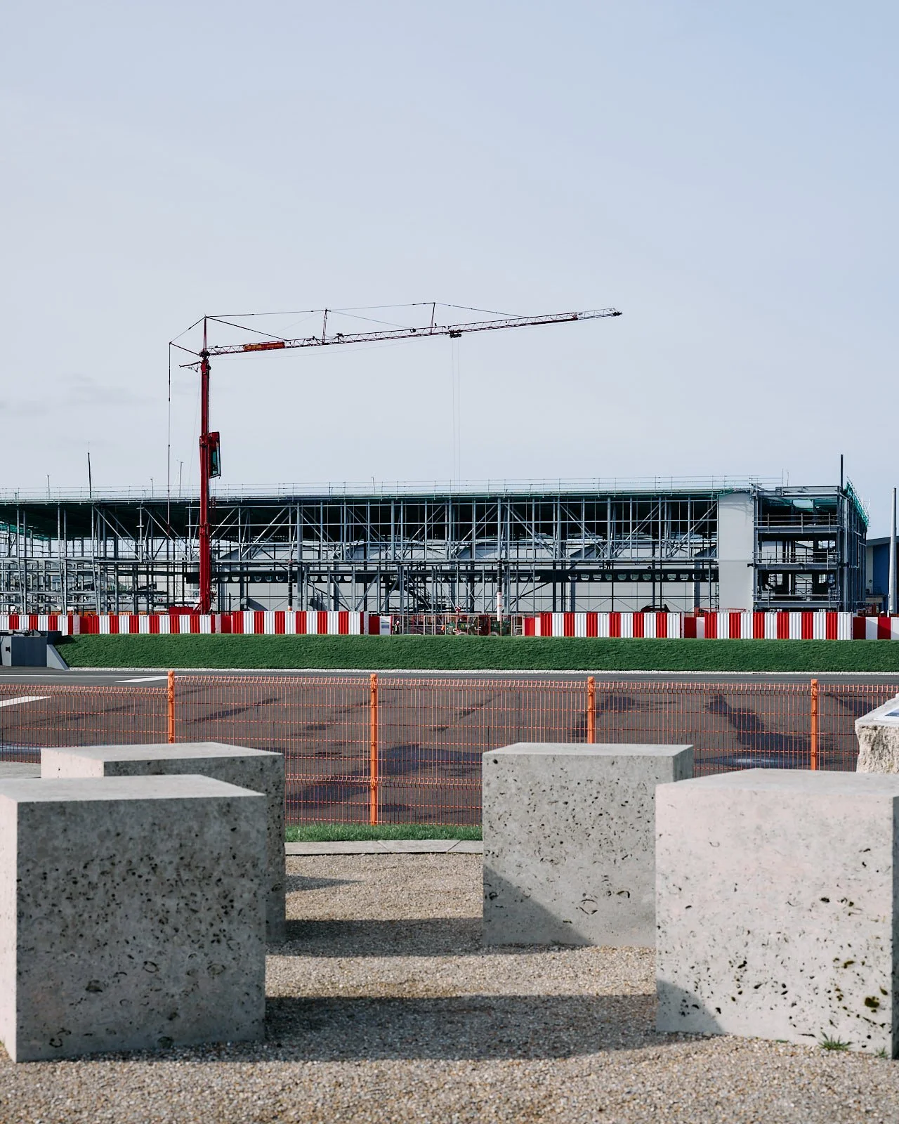 Construction site with a building framework and a tower crane, surrounded by orange fencing and concrete blocks in the foreground.