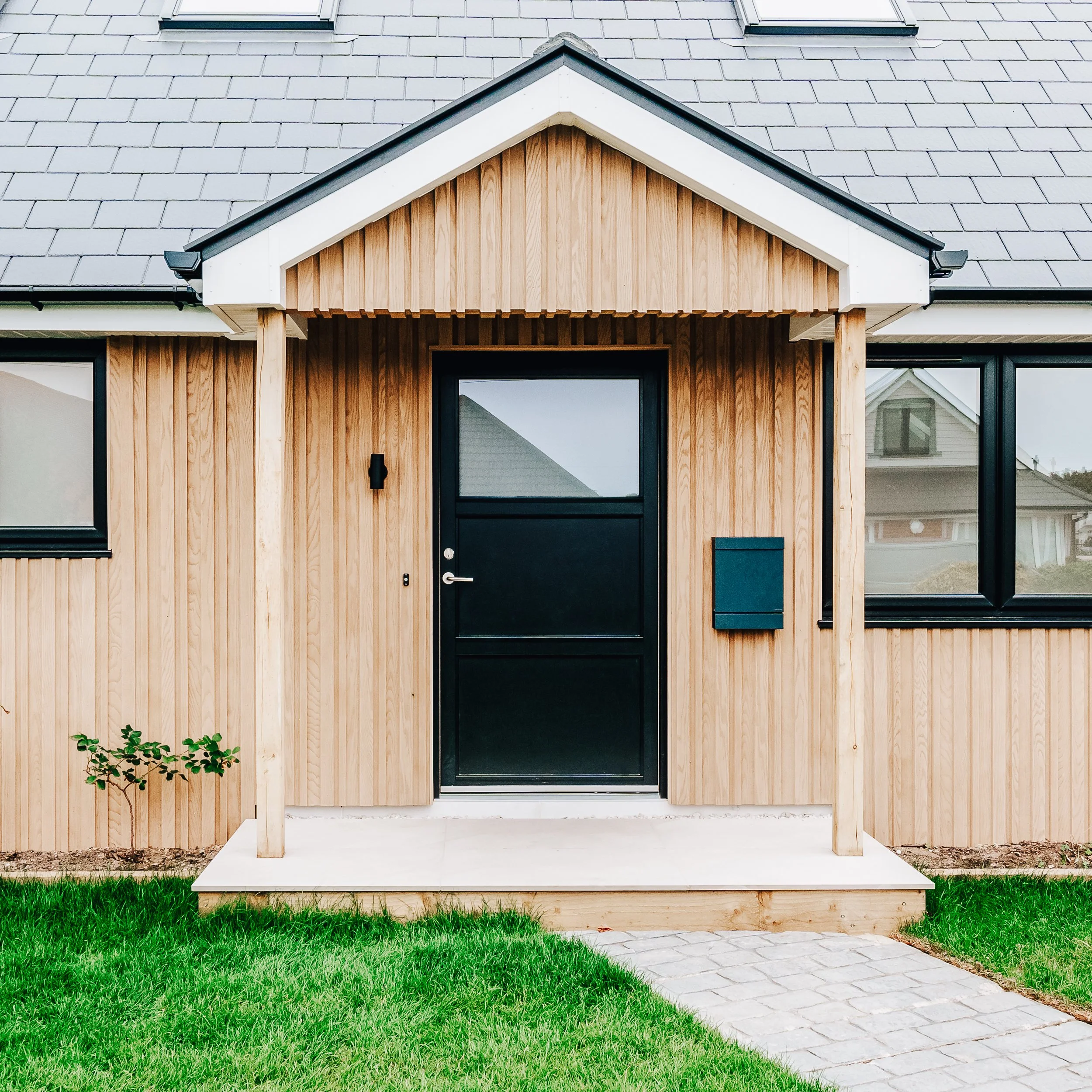 Front entrance of a house with a modern design, featuring a black door, wooden accents, and large windows. The house has a light gray shingled roof with skylights and is surrounded by a green lawn and a paved walkway.