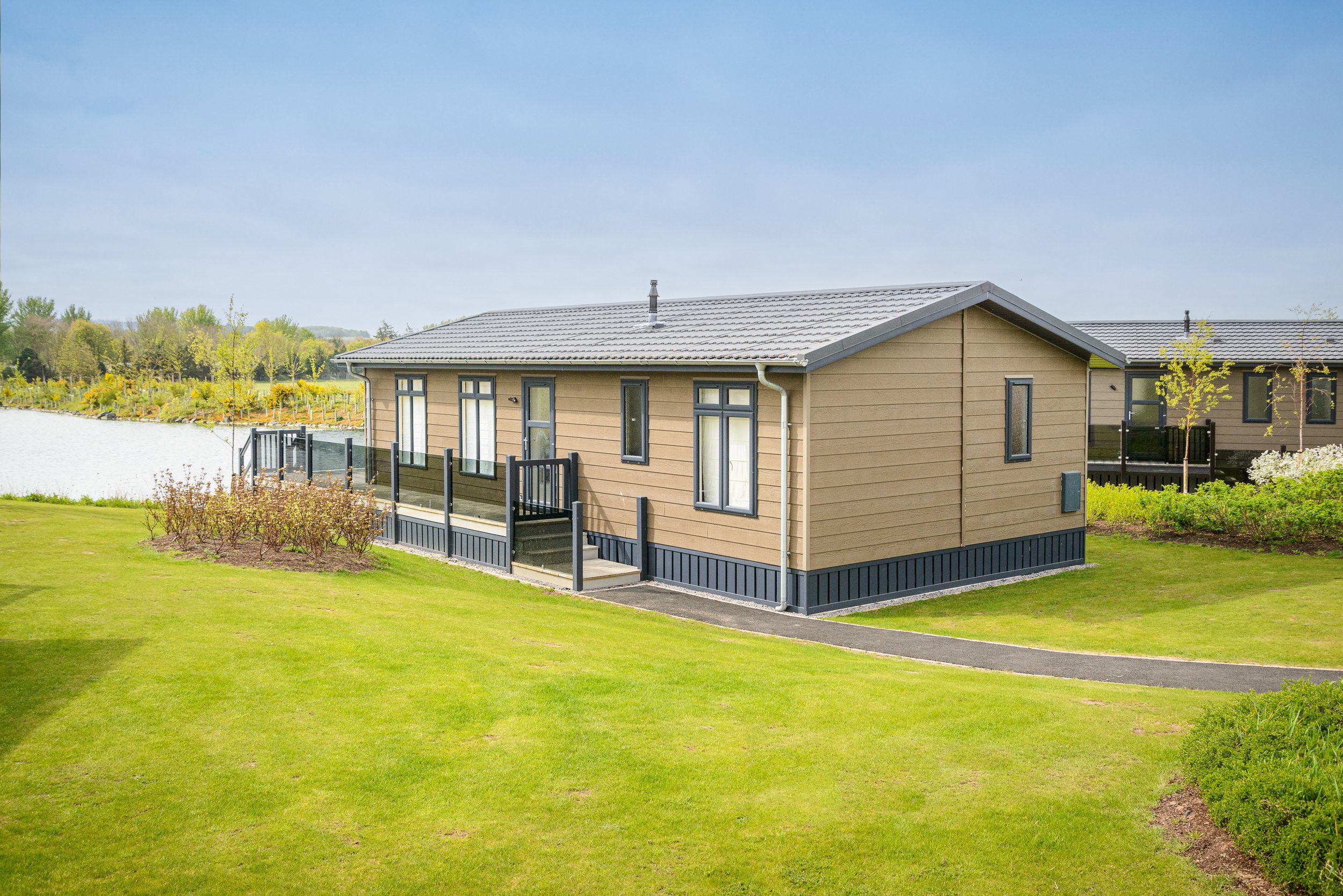 A modern house with beige horizontal siding, black trim around windows, and a gray tiled roof, situated in a lush green landscape near a body of water with trees in the background.