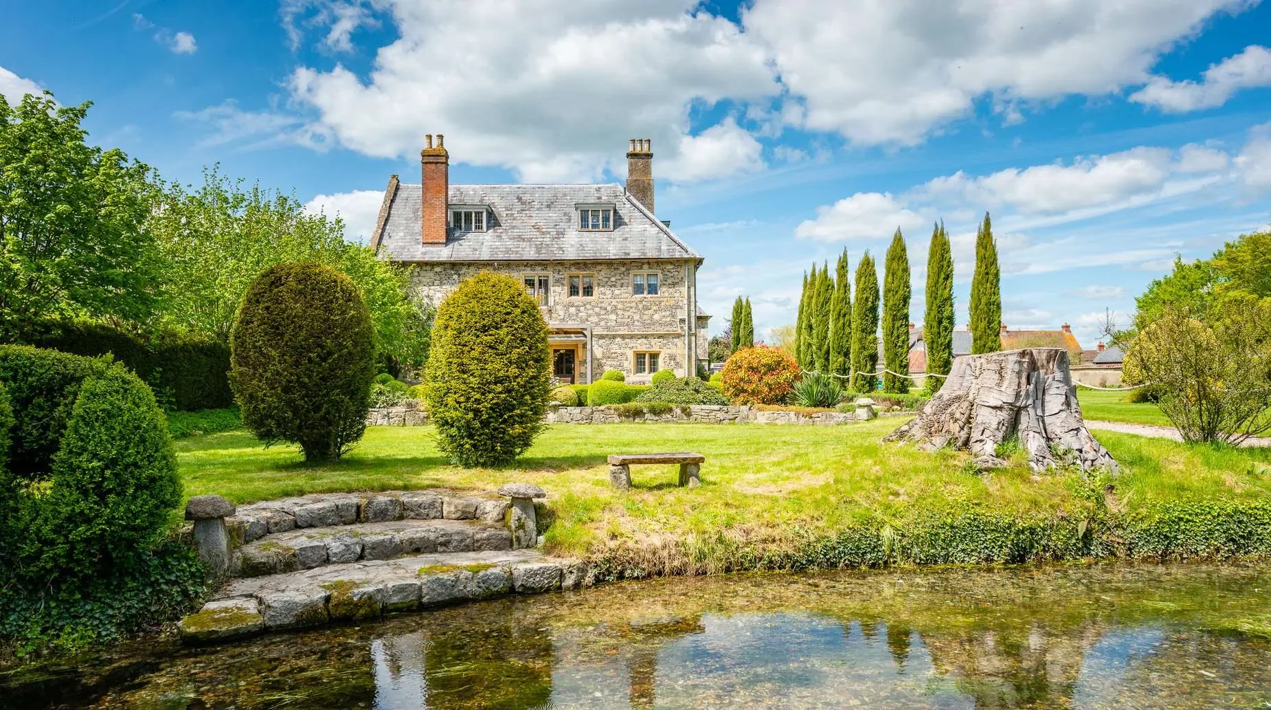 A stone house surrounded by lush greenery, including trees and shrubs, with tall cypress trees in the background, a small pond in the front, and a stone stairway leading to the house, under a partly cloudy blue sky.