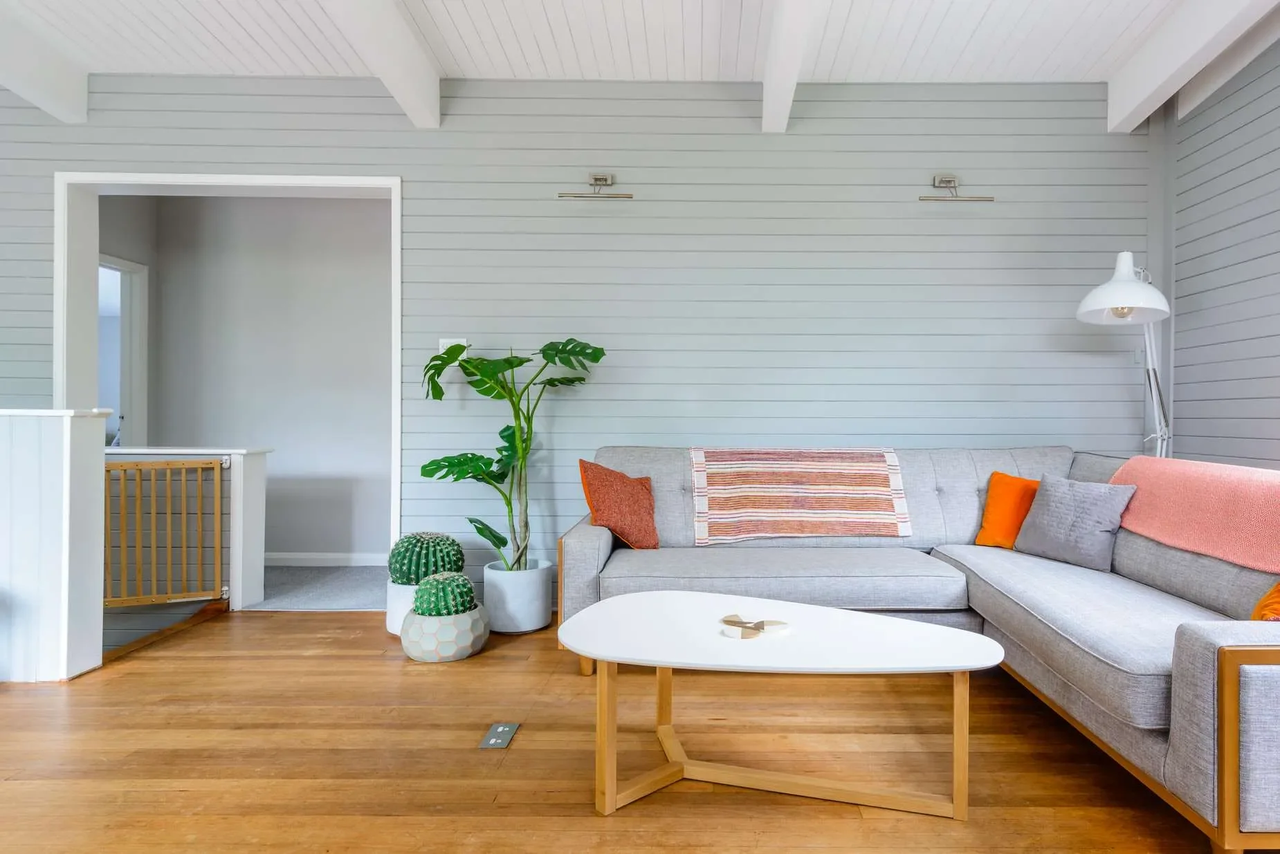 A living room with a sectional sofa, decorative pillows, a white coffee table, houseplants, a wall-mounted lamp, and a white floor lamp, with wood flooring and light gray shiplap walls.