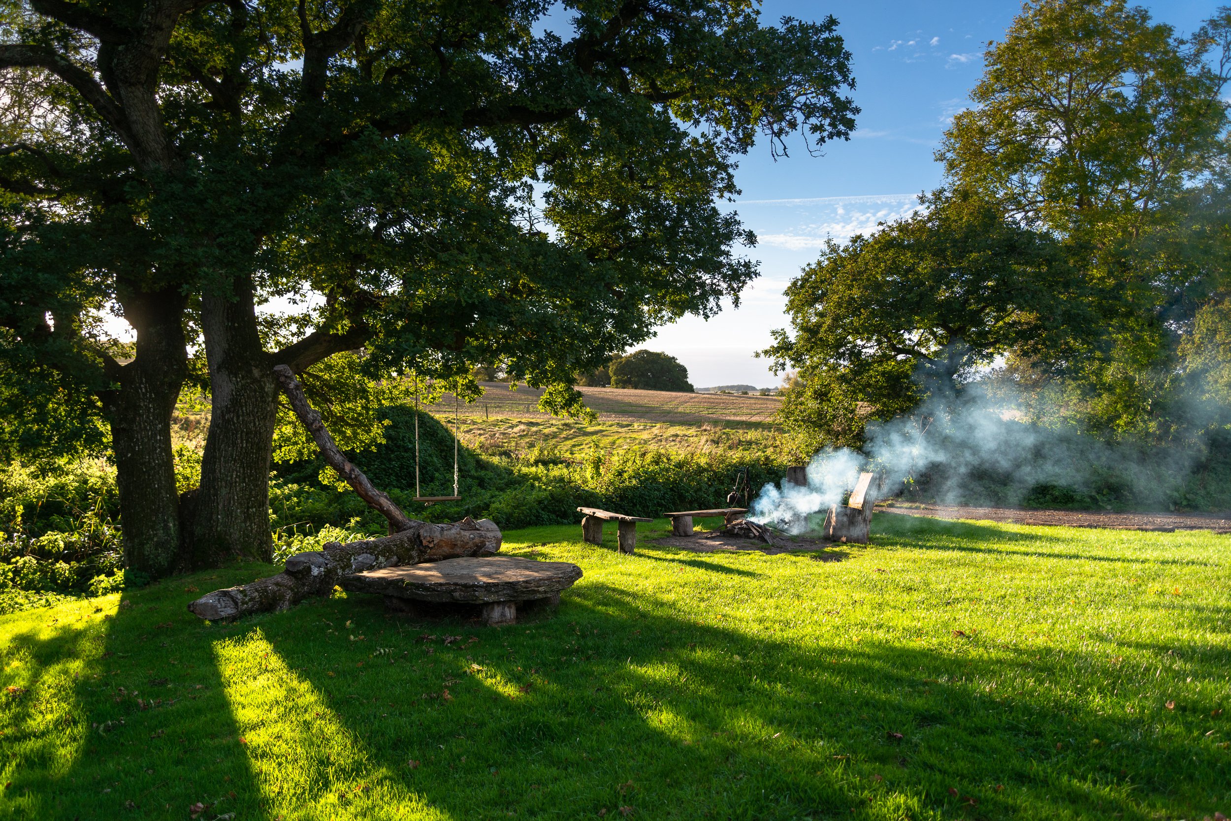 Firepit scene at Stock Gaylard glamping site in Dorset, showing smoke, trees and countryside accommodation in late afternoon light.