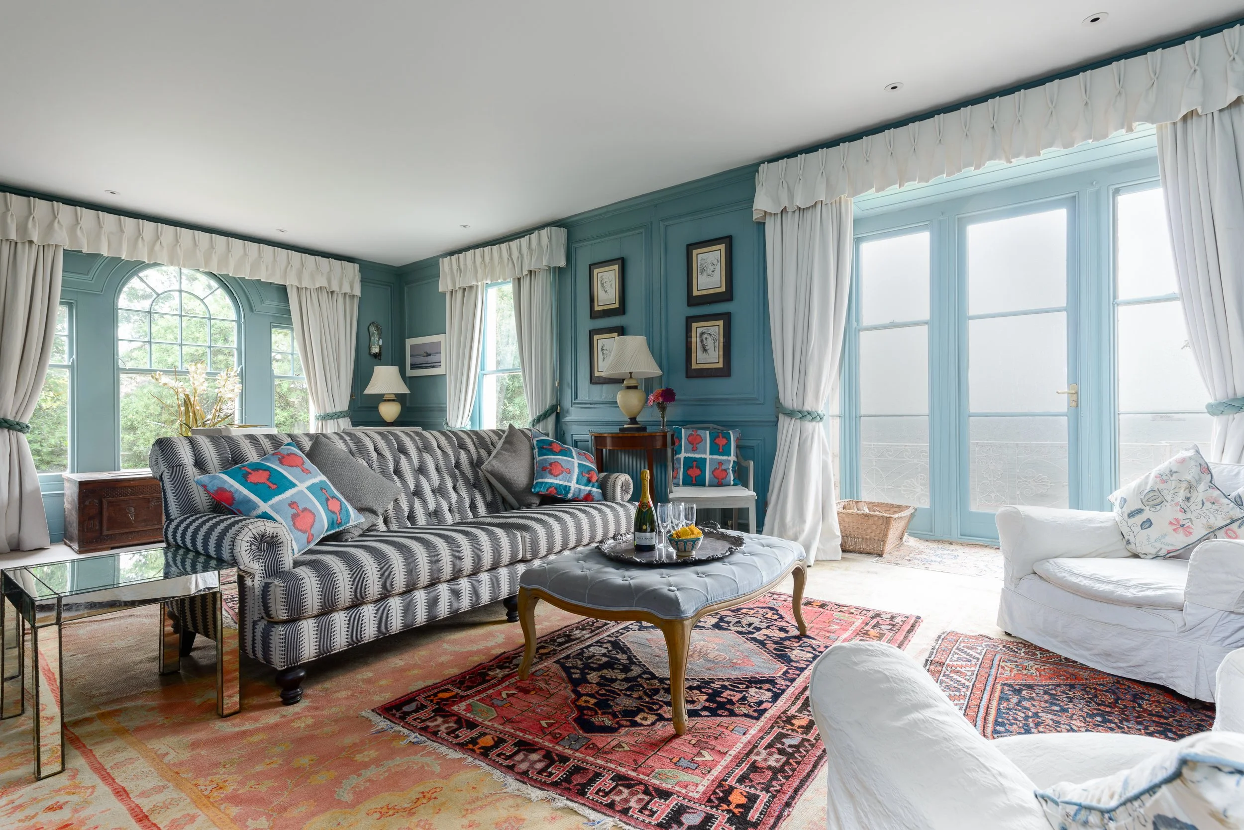 Blue sitting room at The Pink House, showing period-style furnishings, patterned sofa, rugs, large windows and sea views.
