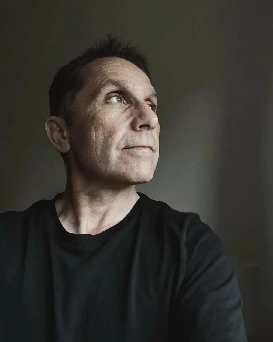 A man with short dark hair and light skin, wearing a black shirt, looking slightly upward and to the side against a plain dark background.