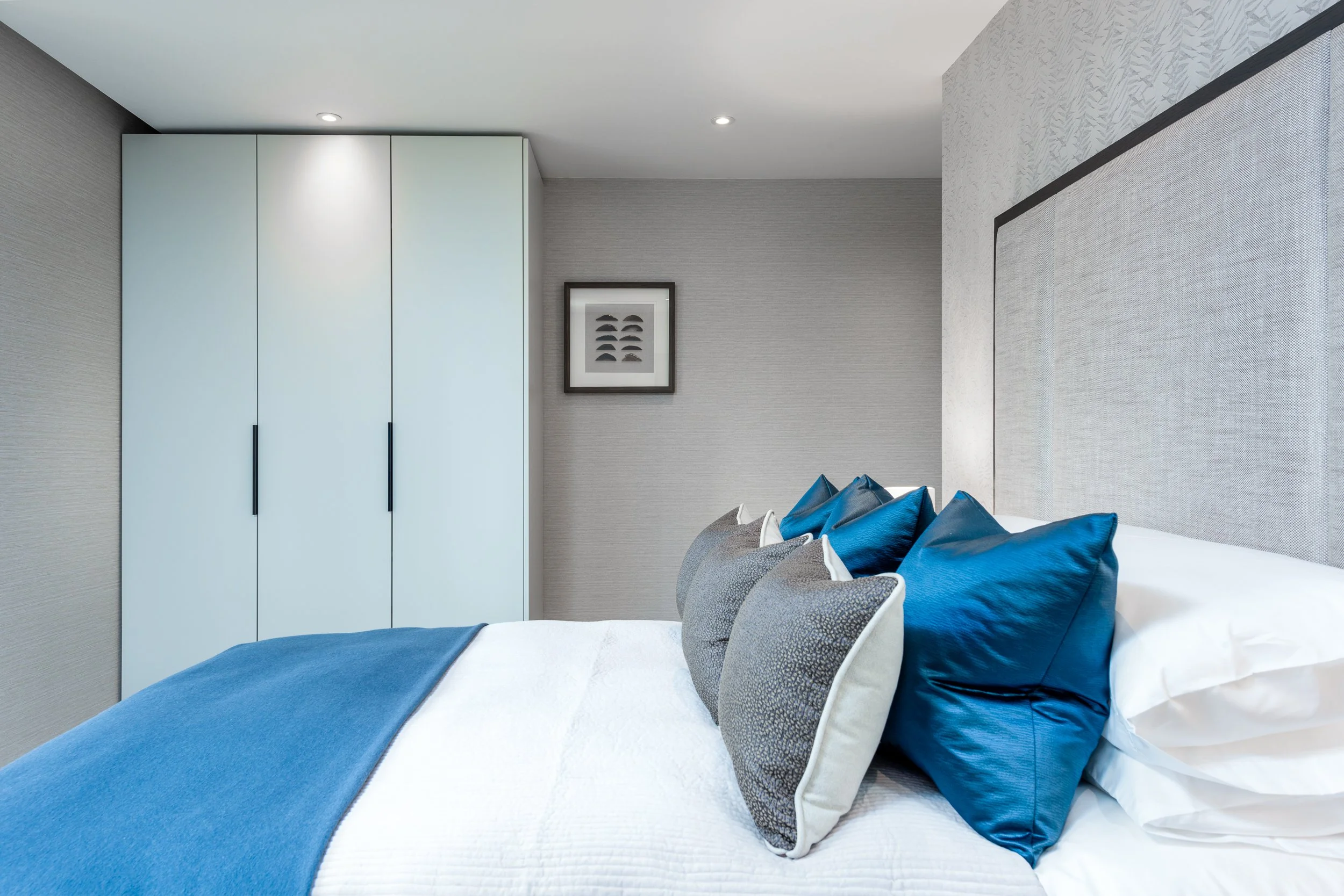 Modern bedroom interior featuring upholstered headboard, blue bedding, fitted wardrobes and a neutral colour palette.