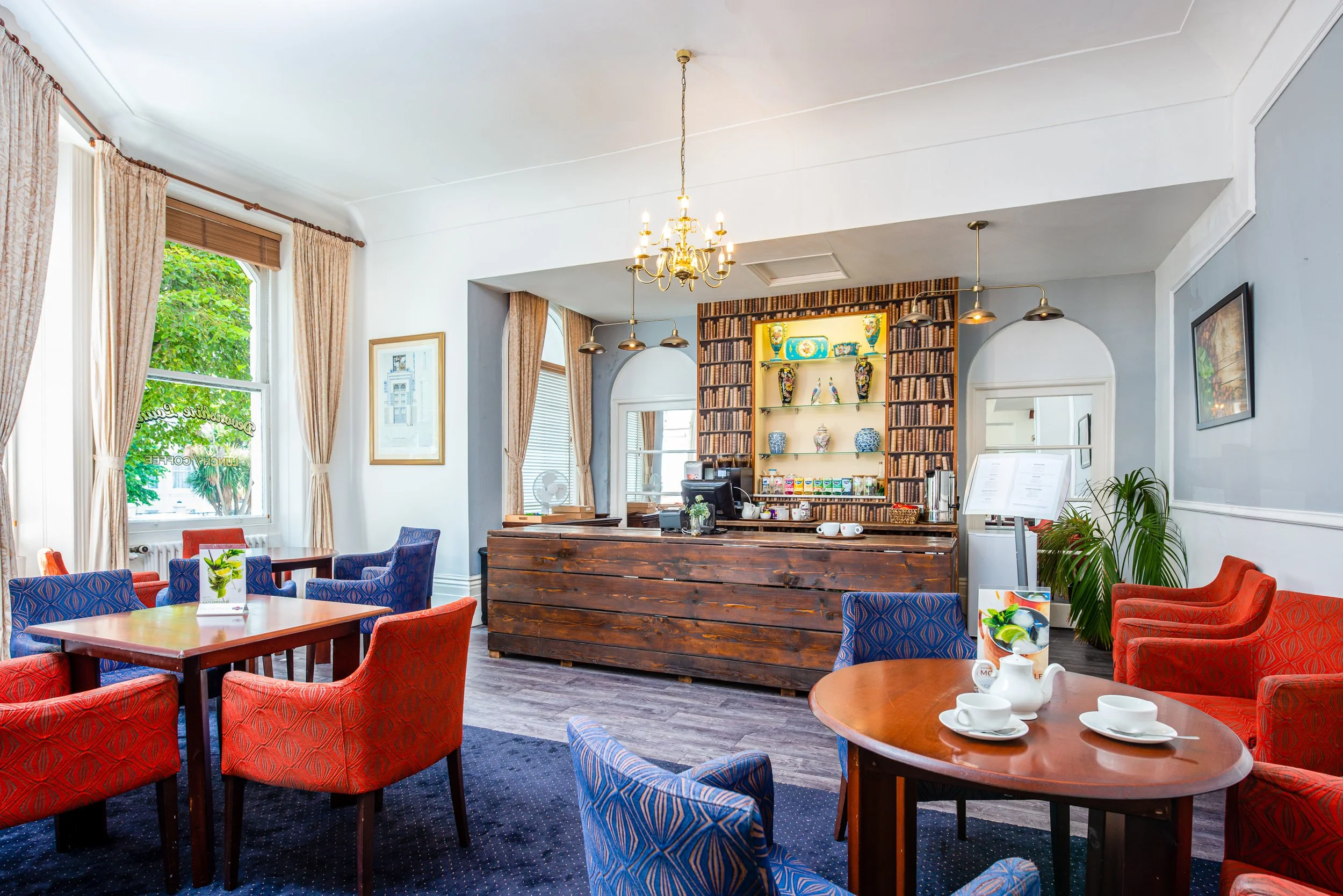 Interior of a hotel with red and blue patterned chairs around wooden tables, large windows with beige curtains, a wooden counter with coffee cups, and a bookshelf decorated with colorful vases and books, illuminated by chandeliers and pendant lights.