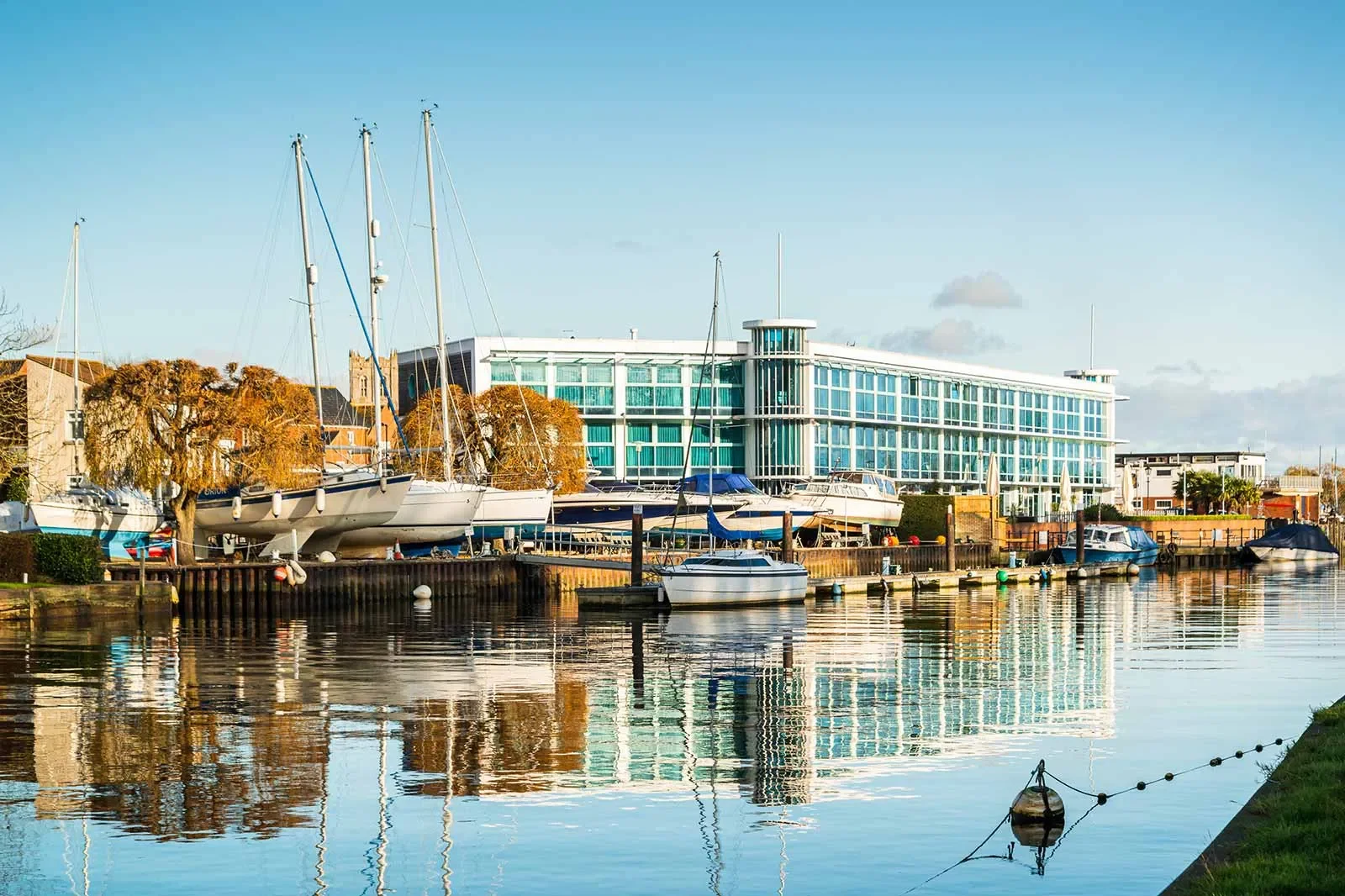 A harbor with sailboats and motorboats docked along the water, modern glass building in the background, trees with autumn foliage, calm water reflecting the scene.