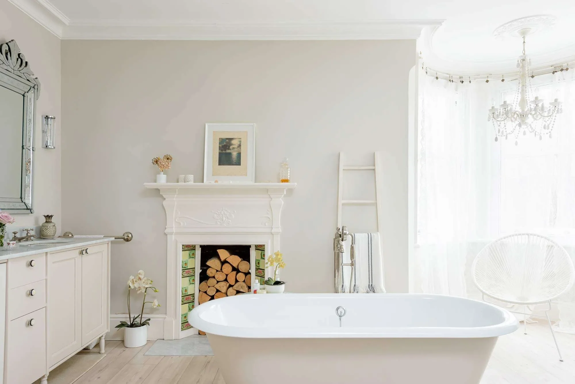 A bright, white bathroom with a freestanding bathtub, a decorative fireplace filled with firewood, a white cabinet with drawers, and a large window with sheer curtains, a chandelier hanging from the ceiling.