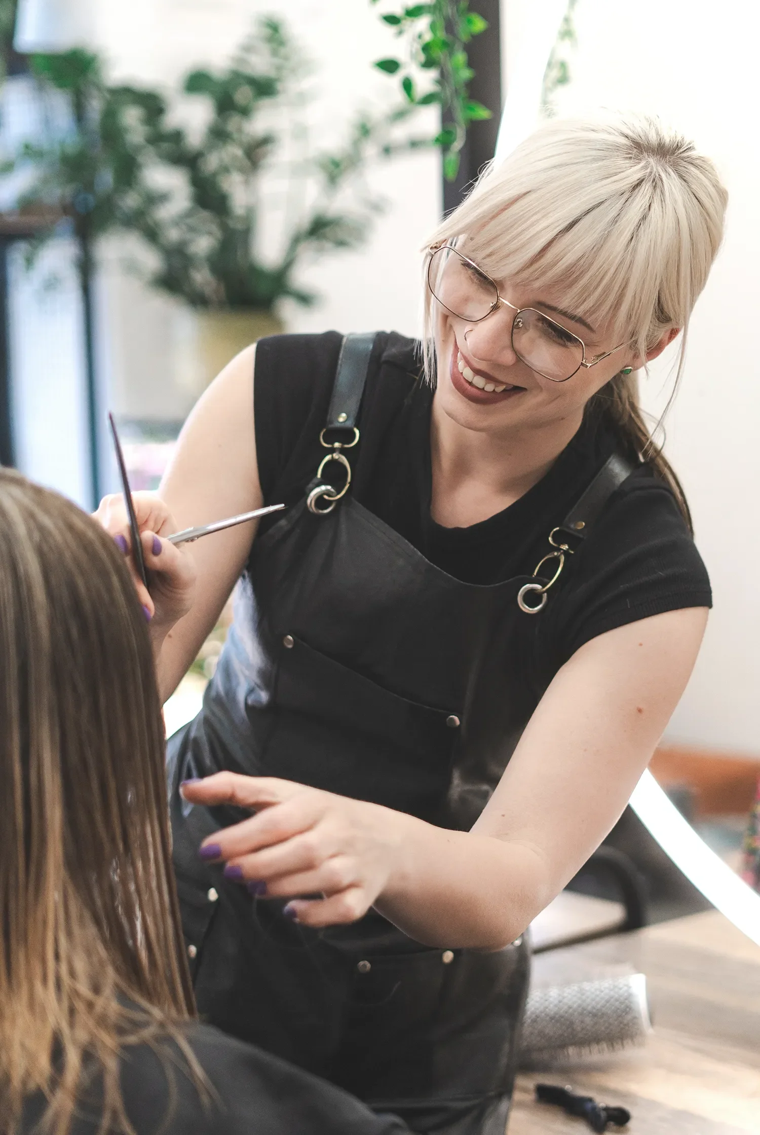 A hairstylist with blonde hair and glasses smiling while working on a client's hair in a salon.